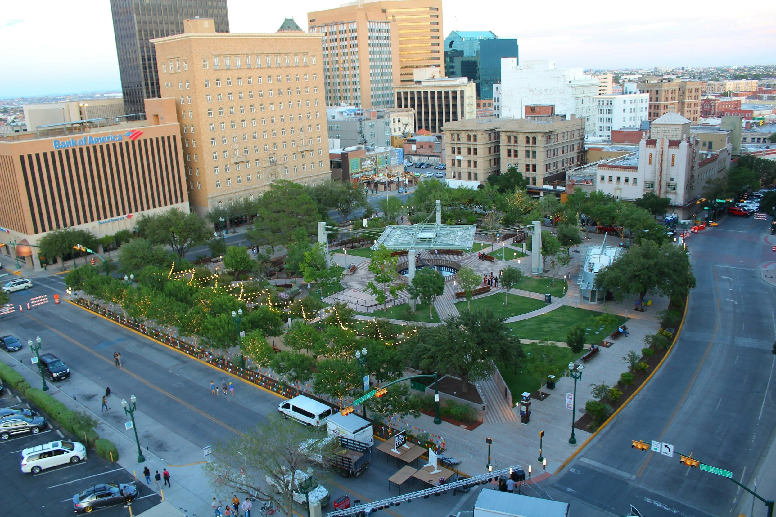 A city square park surrounded by tall buildings with a green lawn, trees, pathways, benches, and string lights. There are people walking and sitting, with cars parked along the streets and some driving by.