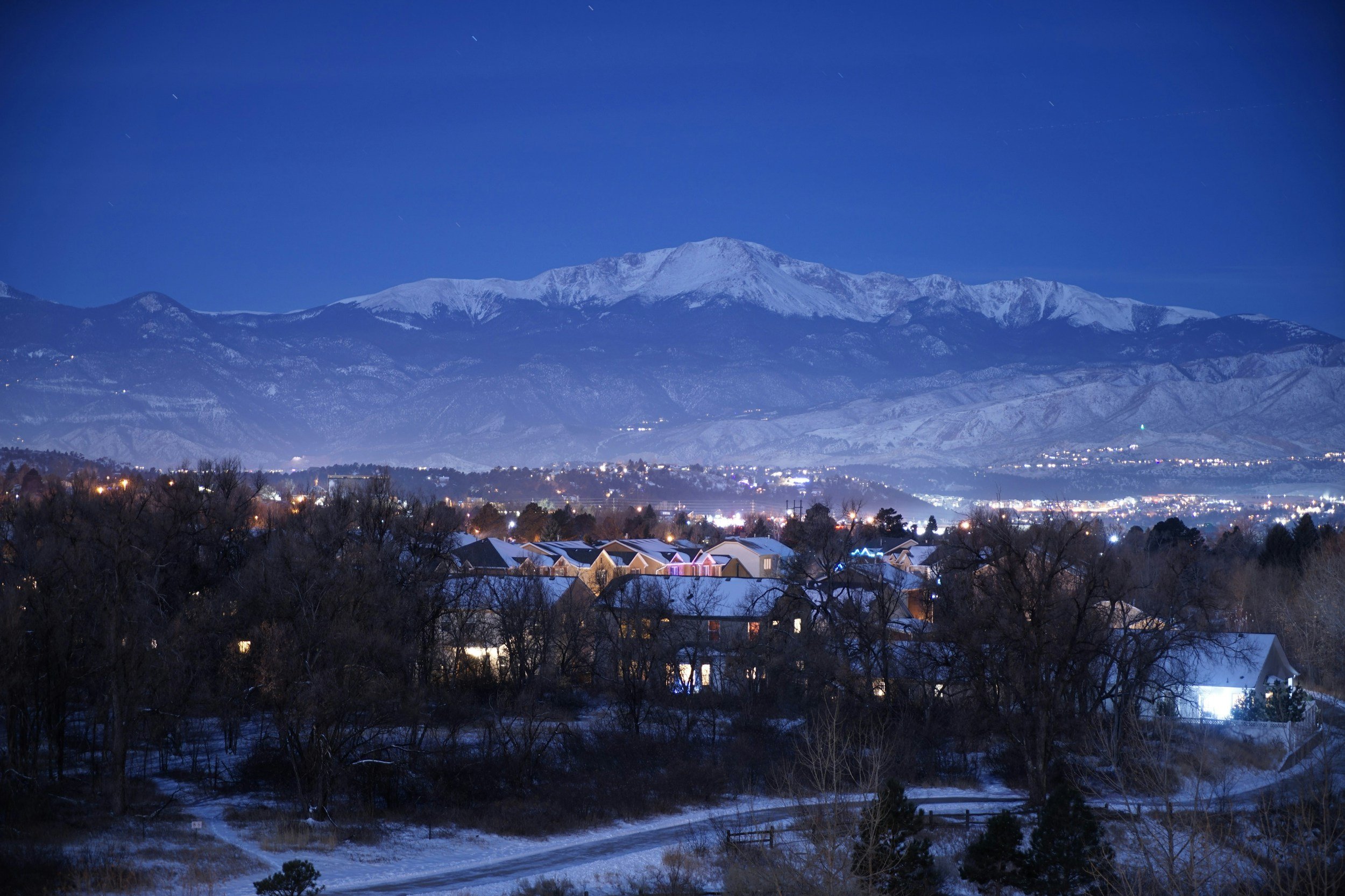 Night view of snow-covered houses, leafless trees, illuminated city in the distance, and snow-capped mountains under a clear blue sky.