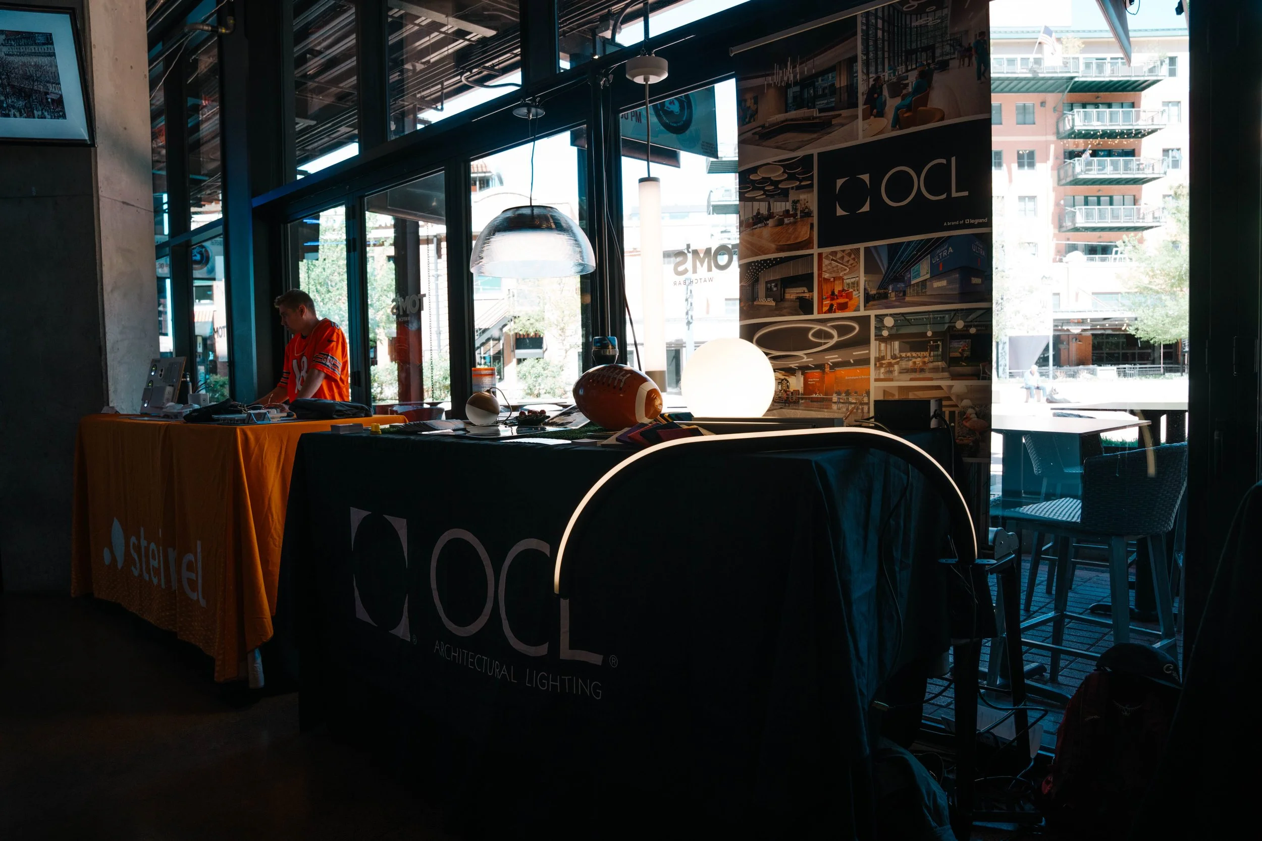 Interior of a modern building with large glass windows, a person wearing an orange sports jersey, a display table with promotional materials, and a wall featuring architectural lighting designs.