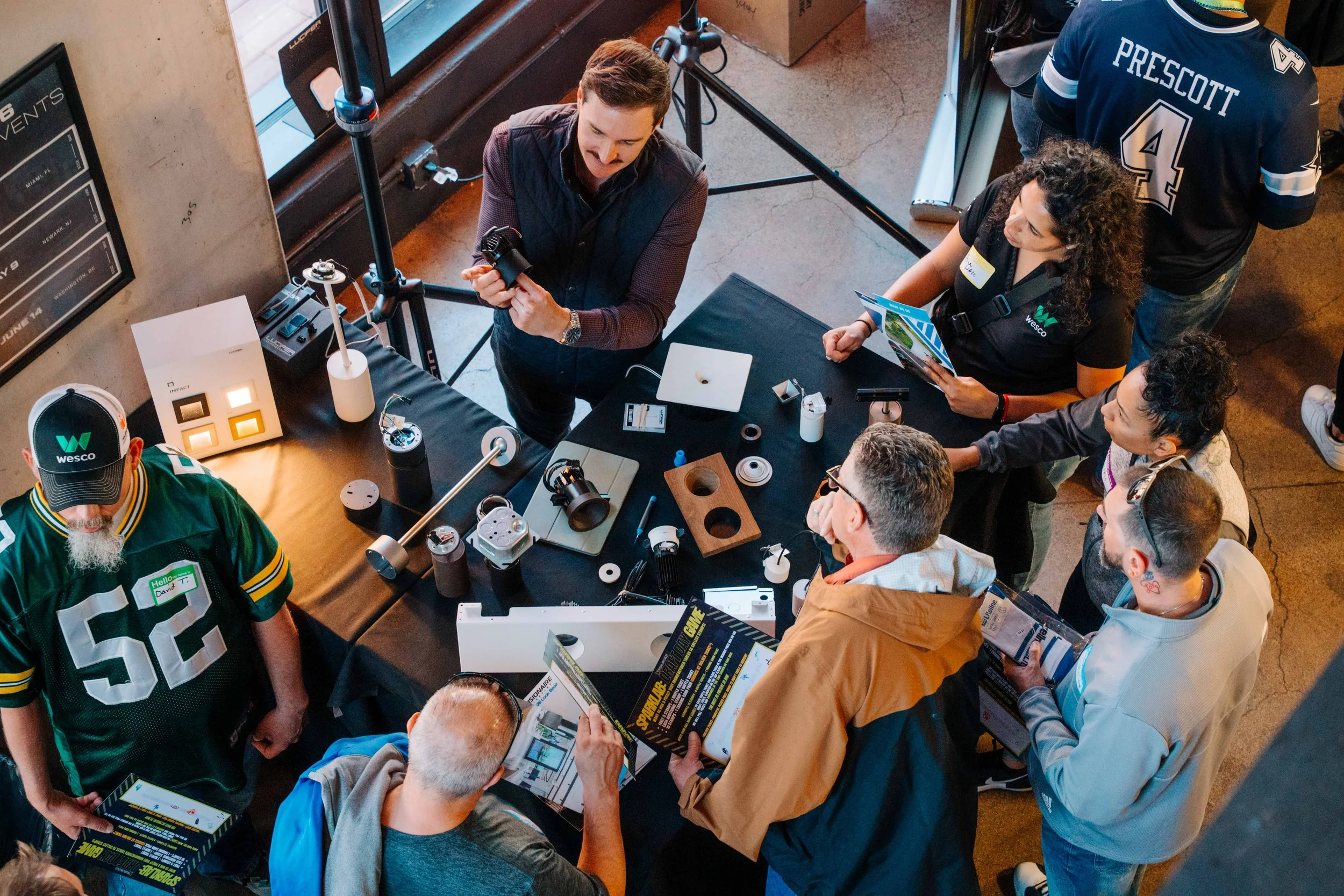 People gathered around a table displaying electronic and technological equipment, engaged in a discussion at an indoor event.