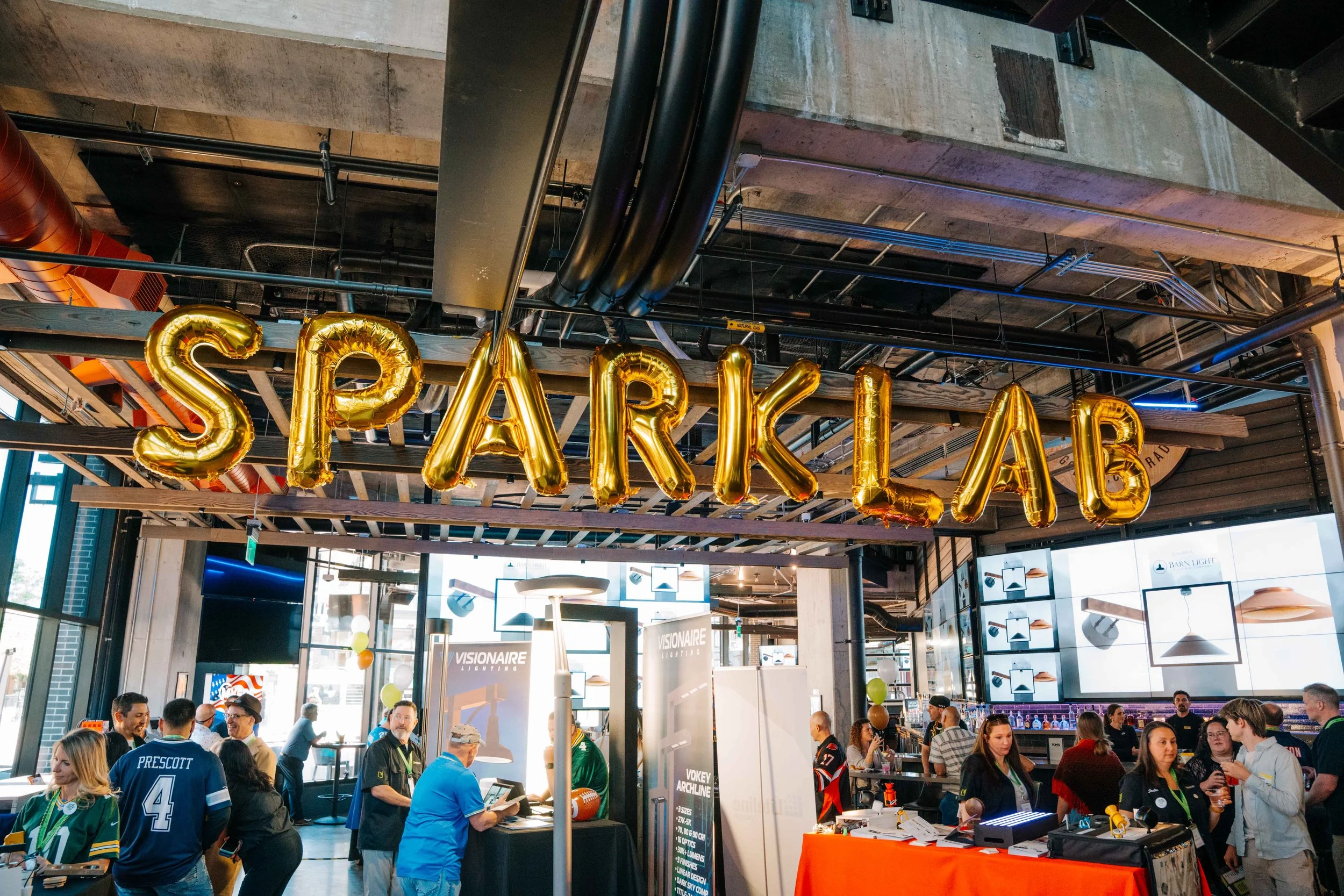 Indoor event with people gathered around booths and displays. Large gold balloons spell out 'SPAR' hanging from the ceiling. The space has an industrial look with exposed pipes and concrete ceiling.