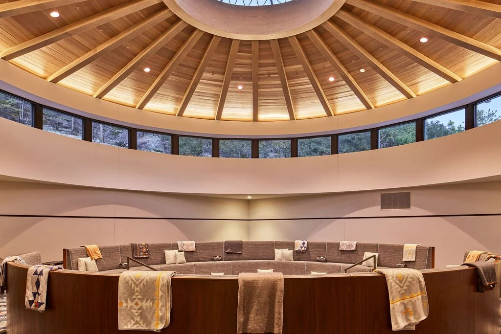 Interior of a modern circular chapel or meeting room with a wooden domed ceiling, large windows around the upper wall, and a semi-circular seating area with blankets and pillows.