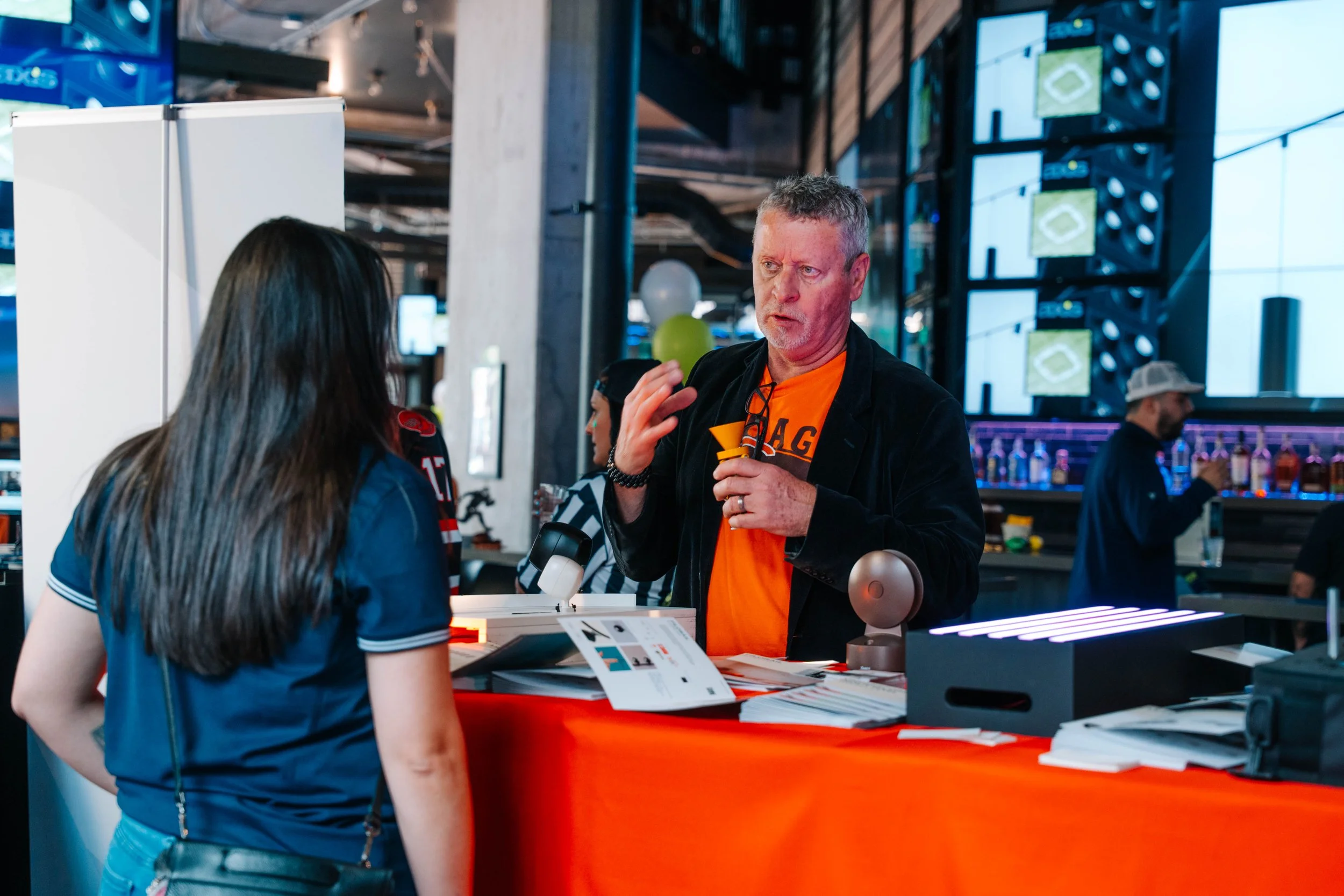 An older man with gray hair and a beard, wearing an orange shirt and black jacket, is speaking to a woman in a sports jersey at a counter in a sports bar or stadium. There are people in the background, TVs displaying a baseball game, and various item