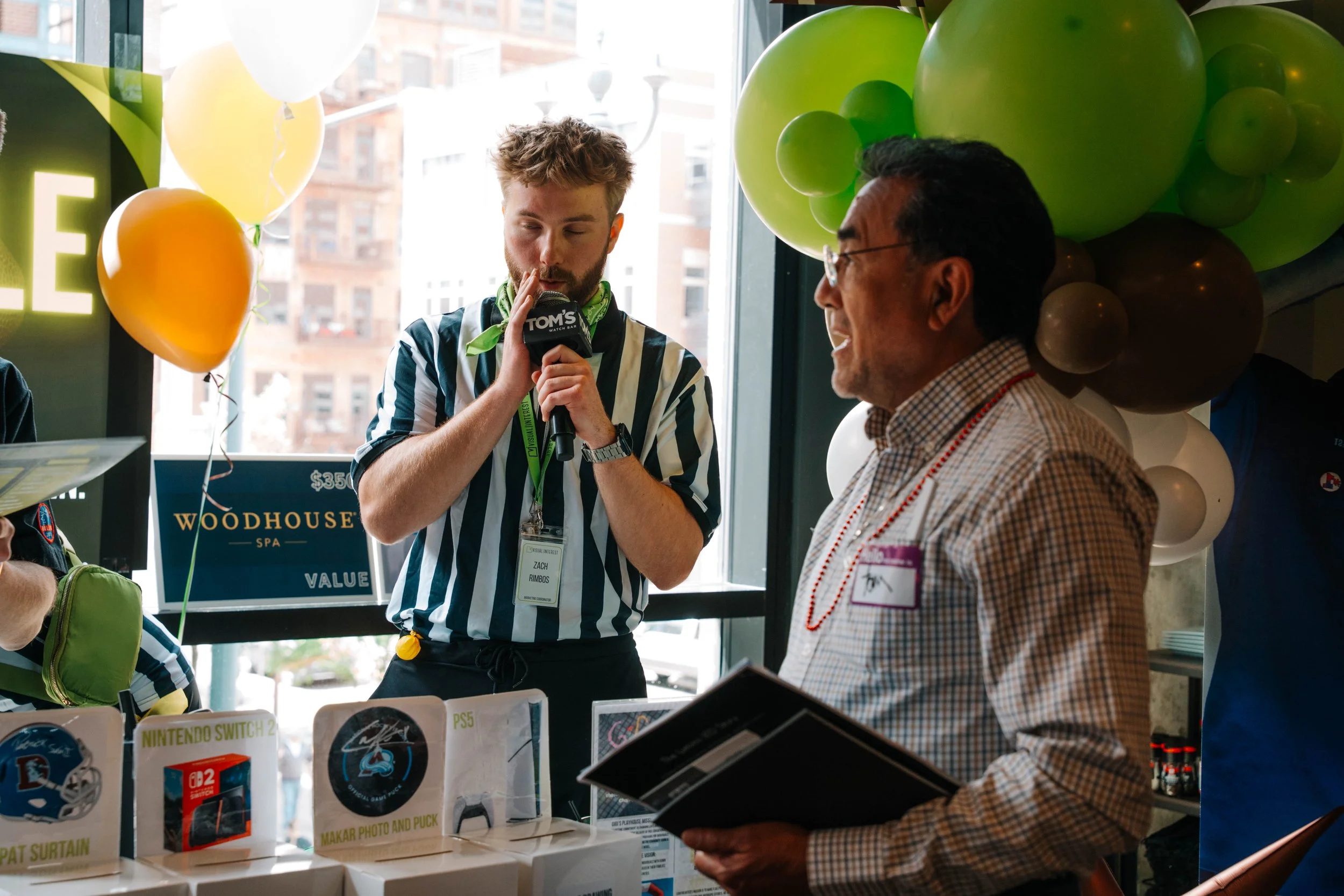 A young man with a beard and a green bandana around his neck speaking into a microphone at an event, standing next to an older man wearing glasses and a checked shirt. There are green, yellow, and white balloons in the background and a table with vid