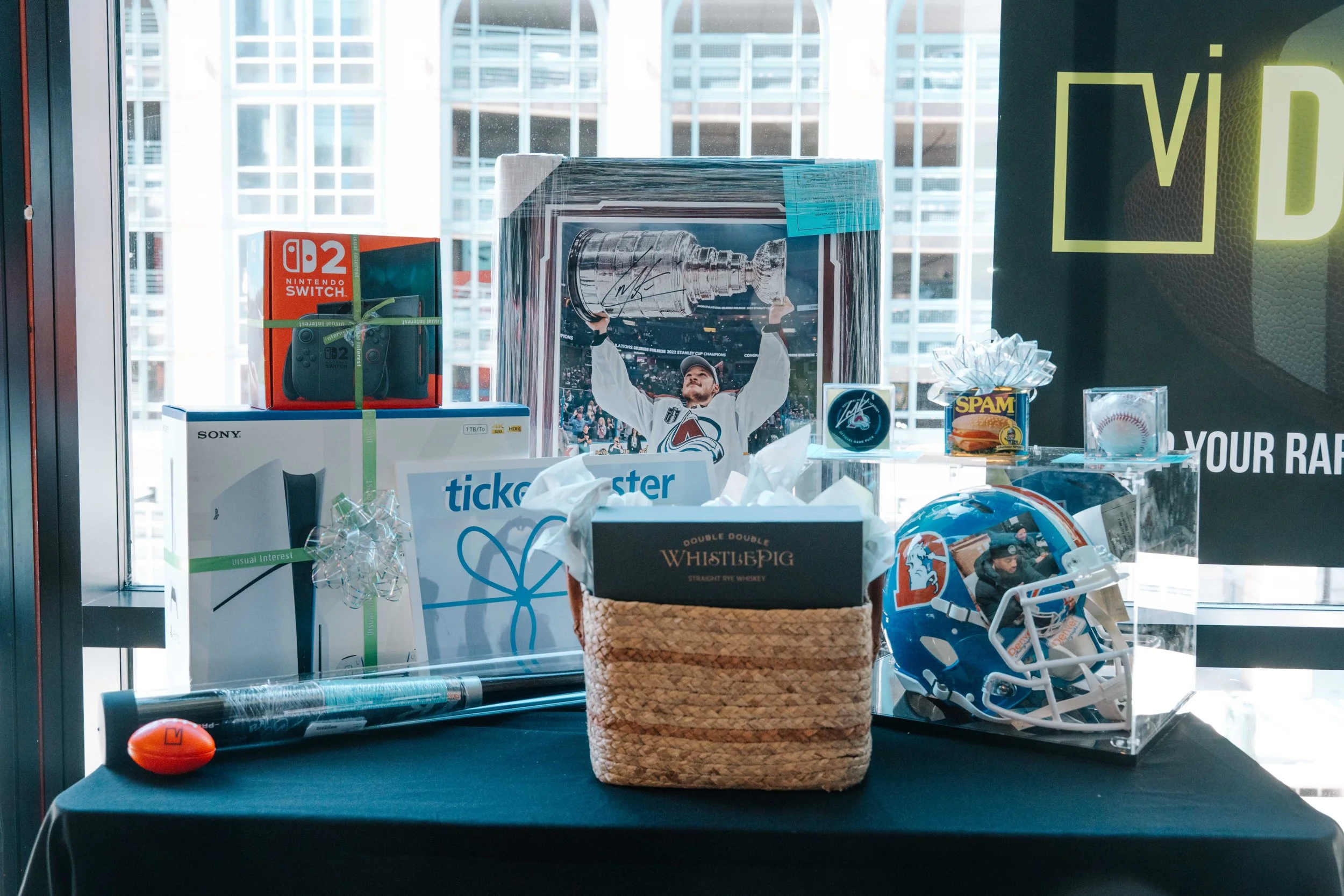 Display of various items including a framed hockey player photograph, a Nintendo Switch box, a whiskey basket, a baseball helmet, and a basketball on a shelf near a window with city buildings in the background.