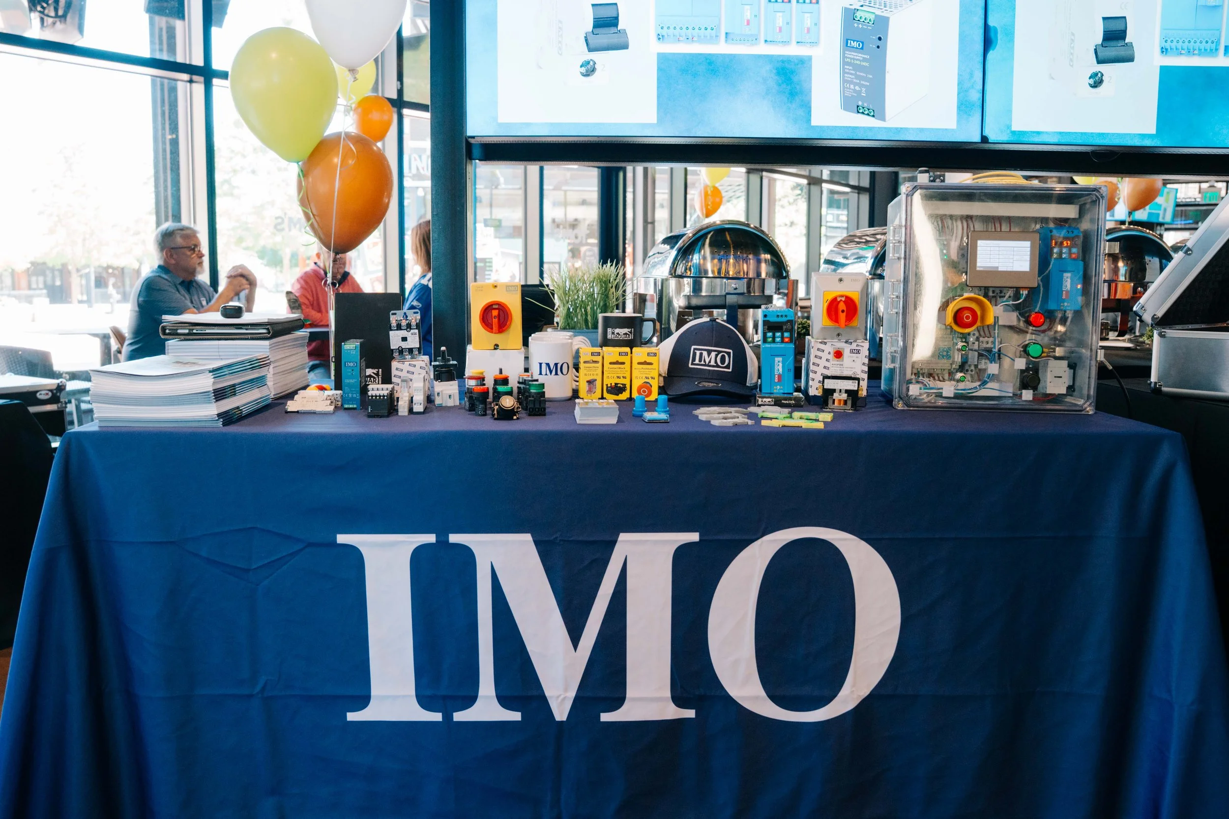 Table covered with various electrical and industrial components, with a blue tablecloth that has the letters 'TMC' printed on it. Behind the table, balloons and people are visible inside a glass-walled building.