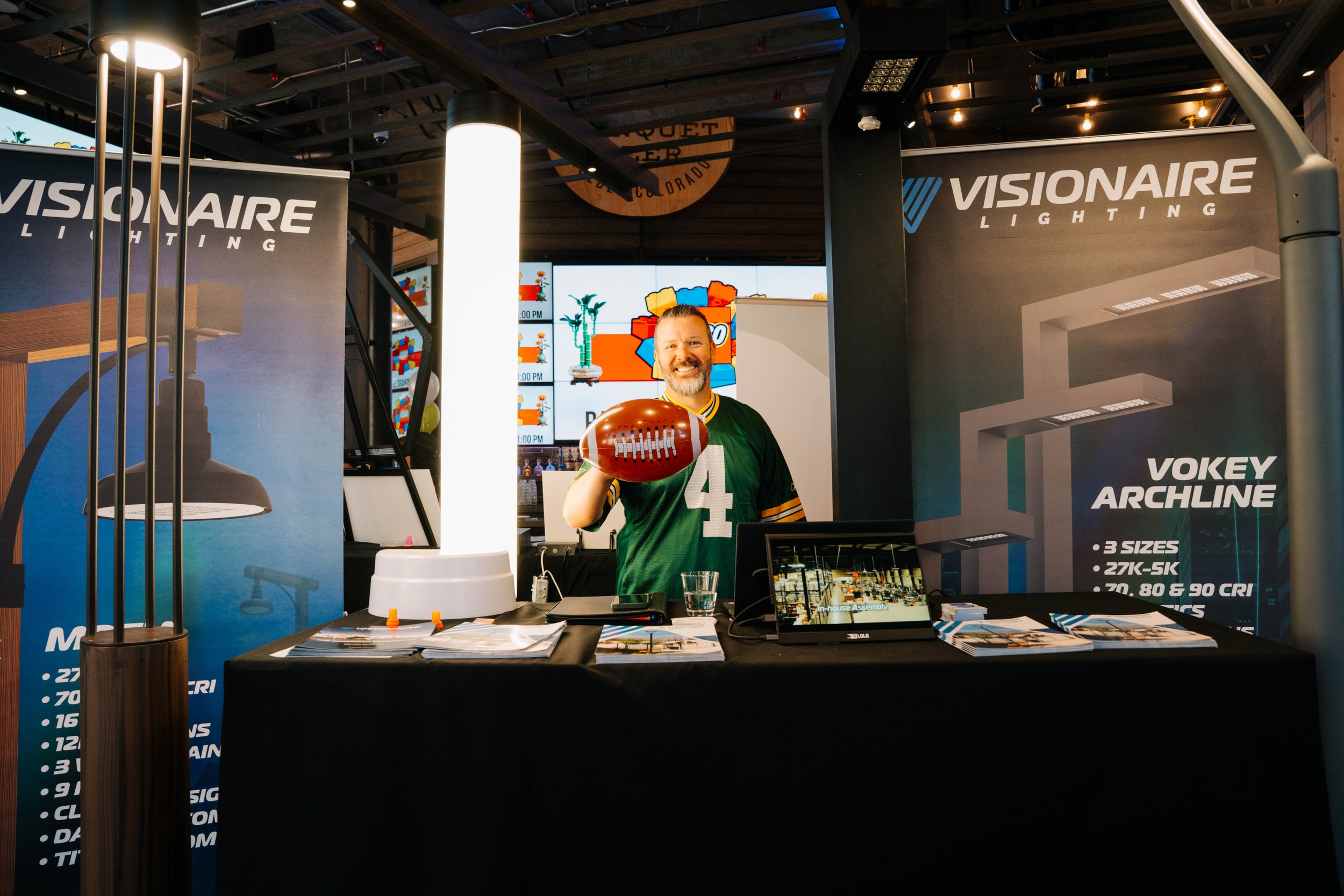 A man with a beard and short hair smiling and holding an American football at a trade show booth for Visionaire Lighting. The booth has banners with product information and a large digital screen in the background displaying animated images.