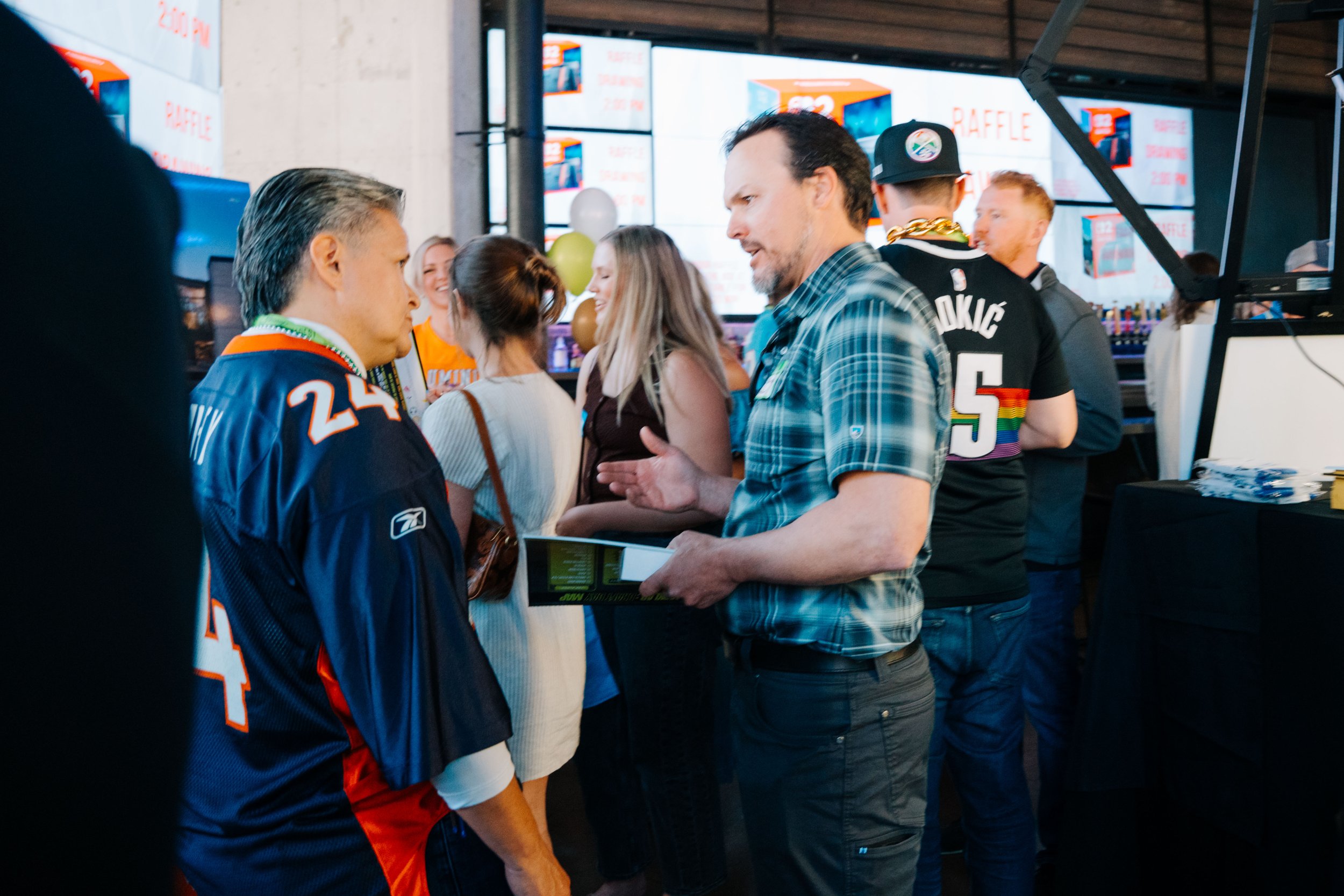 People standing in line at an indoor event, engaging in conversation. Behind them, a large screen displays the words 'RAFFLE' and images of a prize.