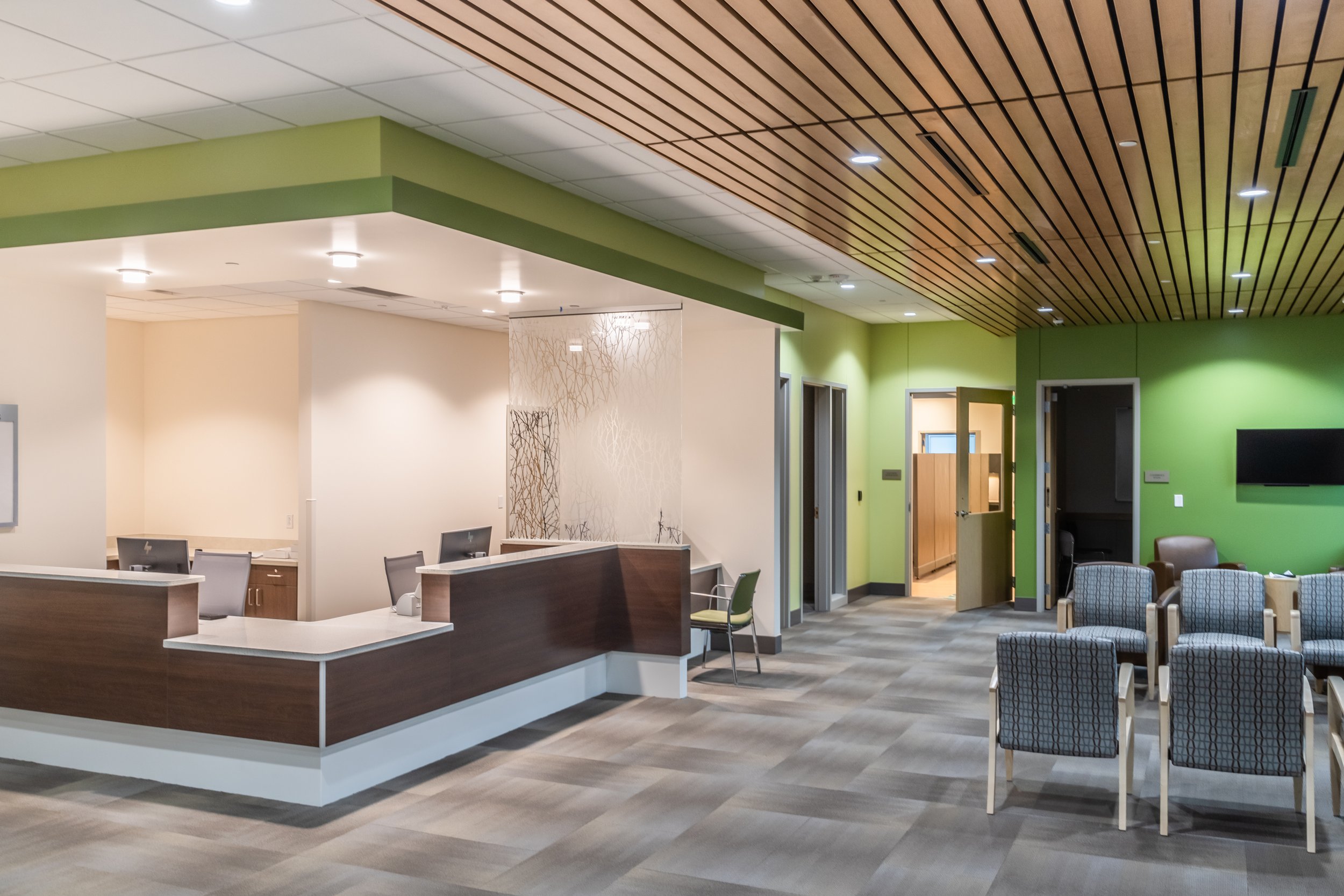 A modern lounge area with various chairs and tables, including green, brown, and blue seating, and a large window to another room with additional seating.