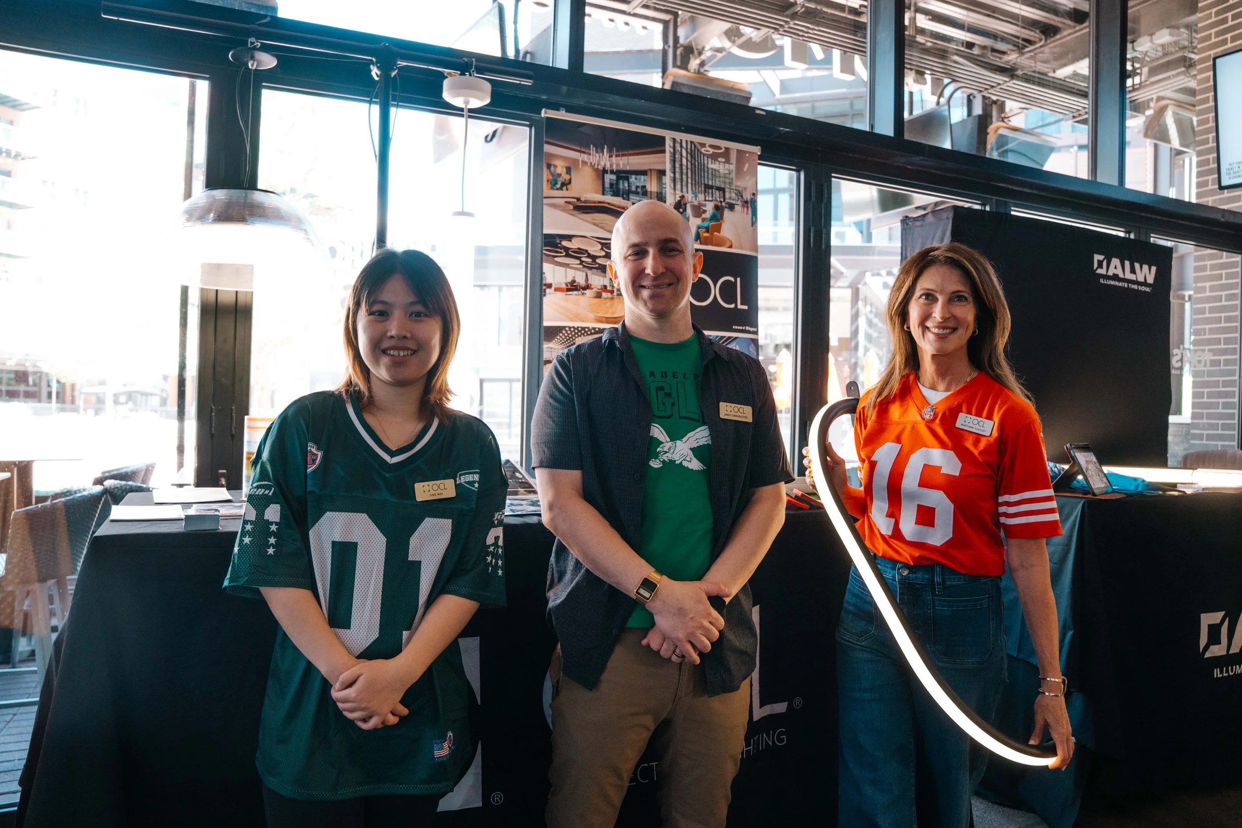 Three people smiling at an indoor event, one woman wearing a green sports jersey, a man in the middle with a blue shirt and a woman on the right in an orange sports jersey, standing near a booth with promotional banners and a ring light.