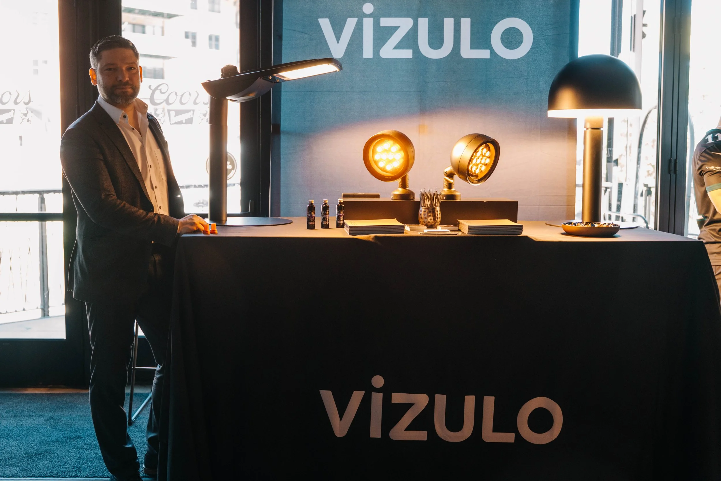 A man in a suit stands at a booth with a blue backdrop displaying the logo 'VIZULO'. The booth has black tablecloth, review lights, and bottles, with sunlight coming through large windows in the background.