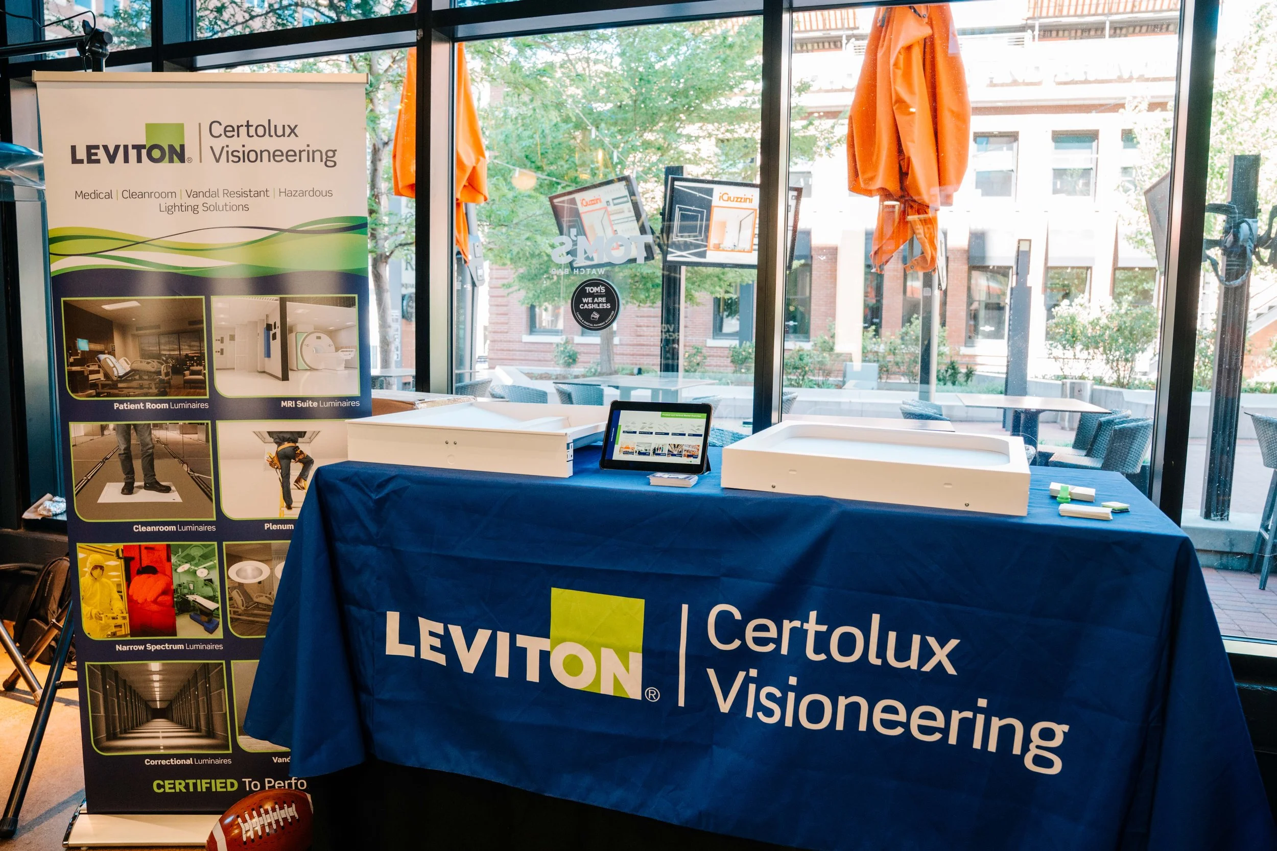 Display booth with a blue tablecloth advertising Leviton Certolux visioning solutions, featuring a promotional poster with images of medical and cleanroom luminaires, and a tablet.