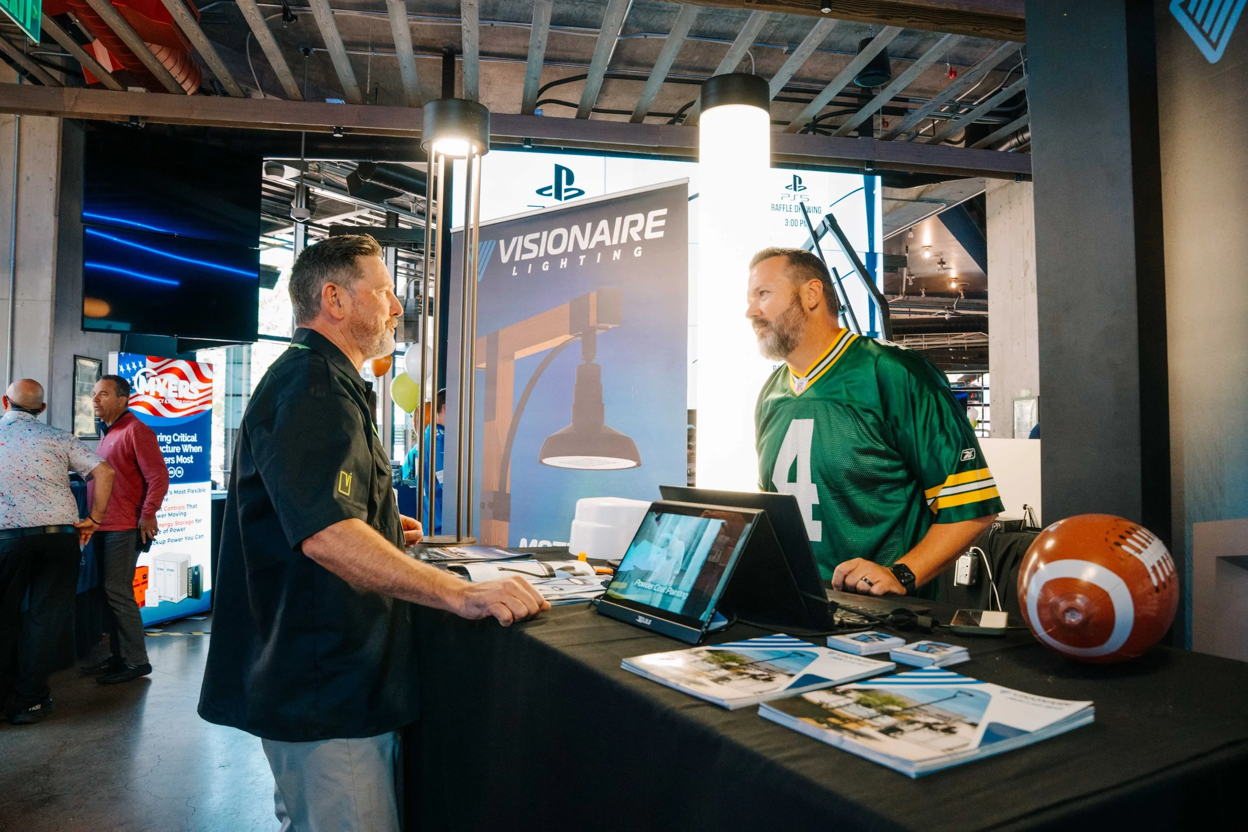 Two men are conversing at a booth, one wearing a black shirt and the other in a green sports jersey, at an indoor event or trade show with banners and displays, including a sign for VISIONAIRE LIGHTING. There are other people in the background, and t
