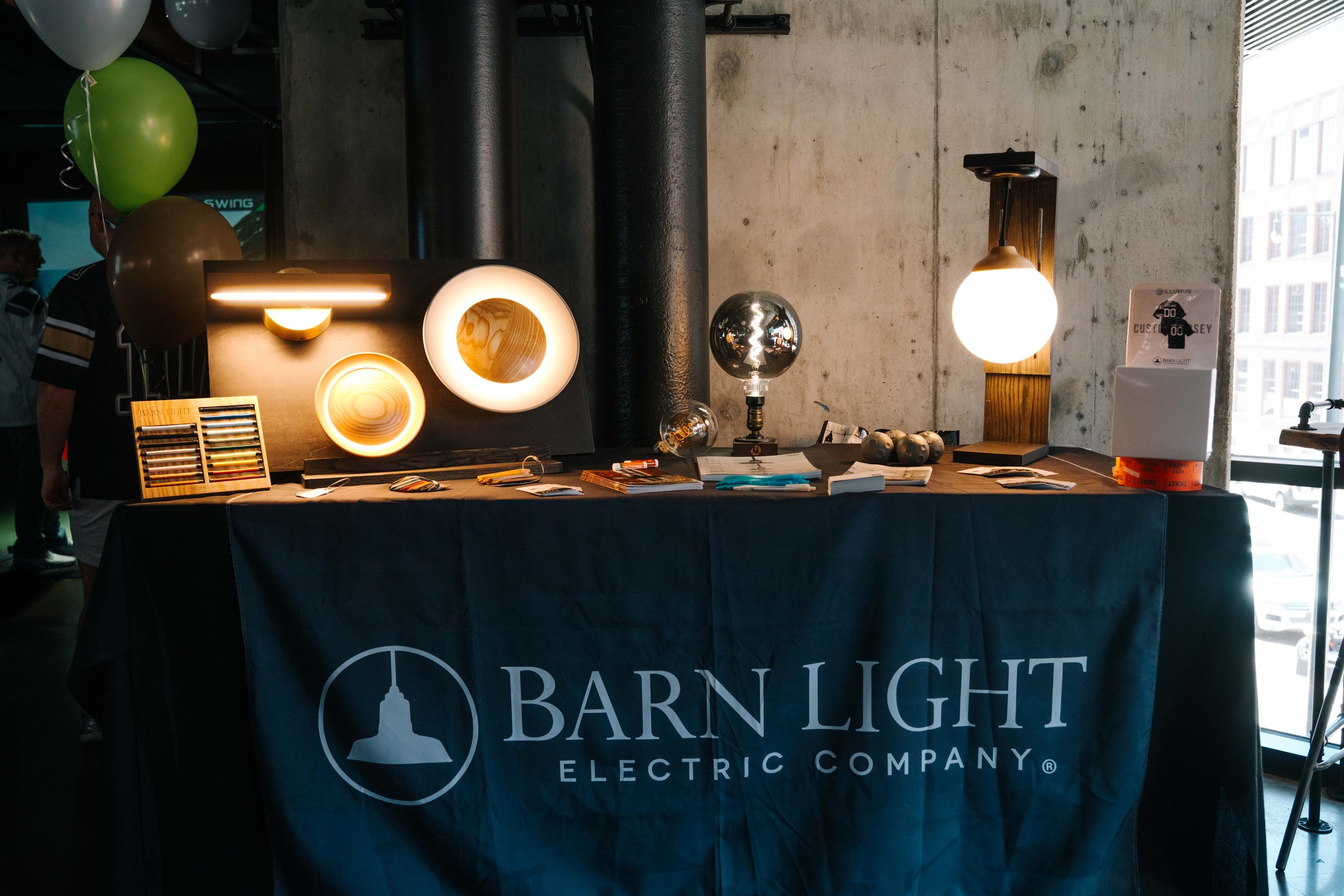 A display table at a trade show features lighting products, including various lamps and bulbs, with a banner reading 'Barn Light Electric Company.' Balloons are attached to the display, and the setting appears to be in a modern industrial space with 