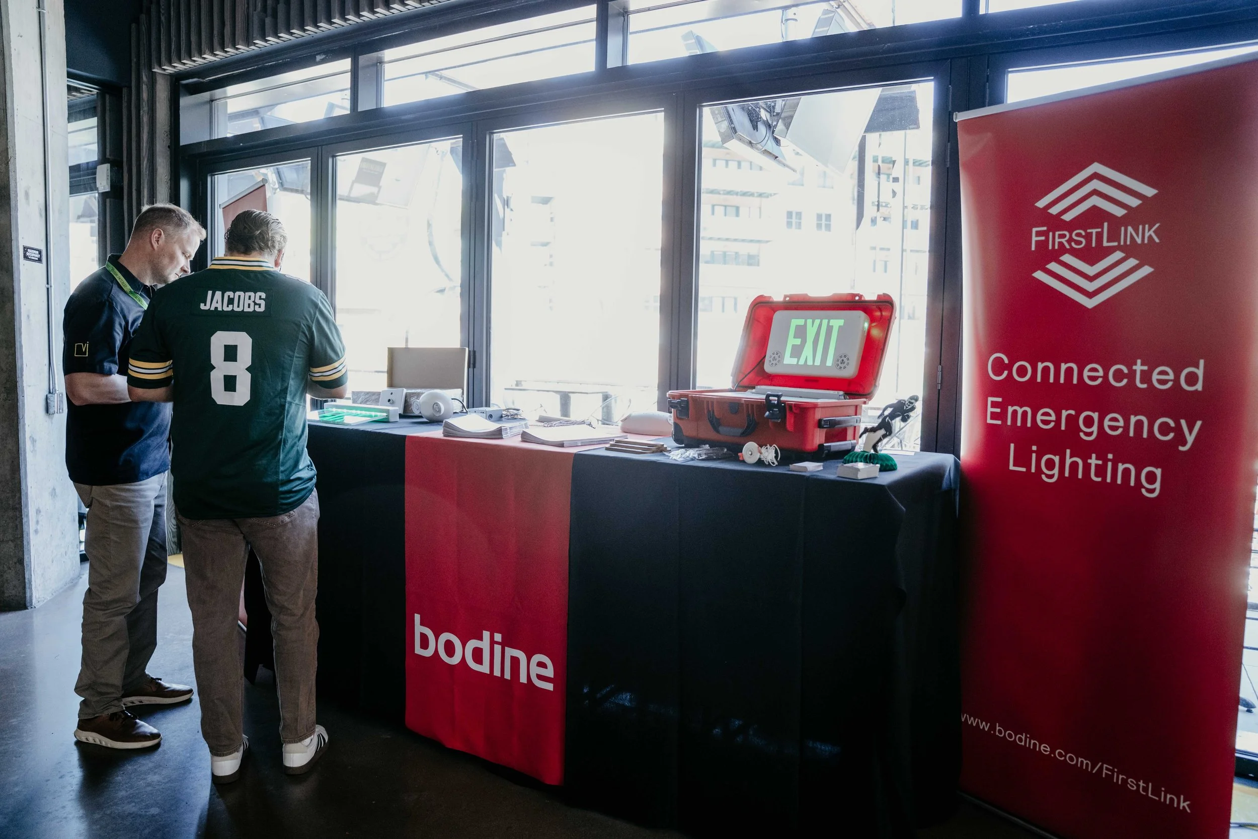 Two men are standing at a table with FirstLink banner and a red emergency lighting box with an illuminated 'EXIT' sign, inside a building with large windows.