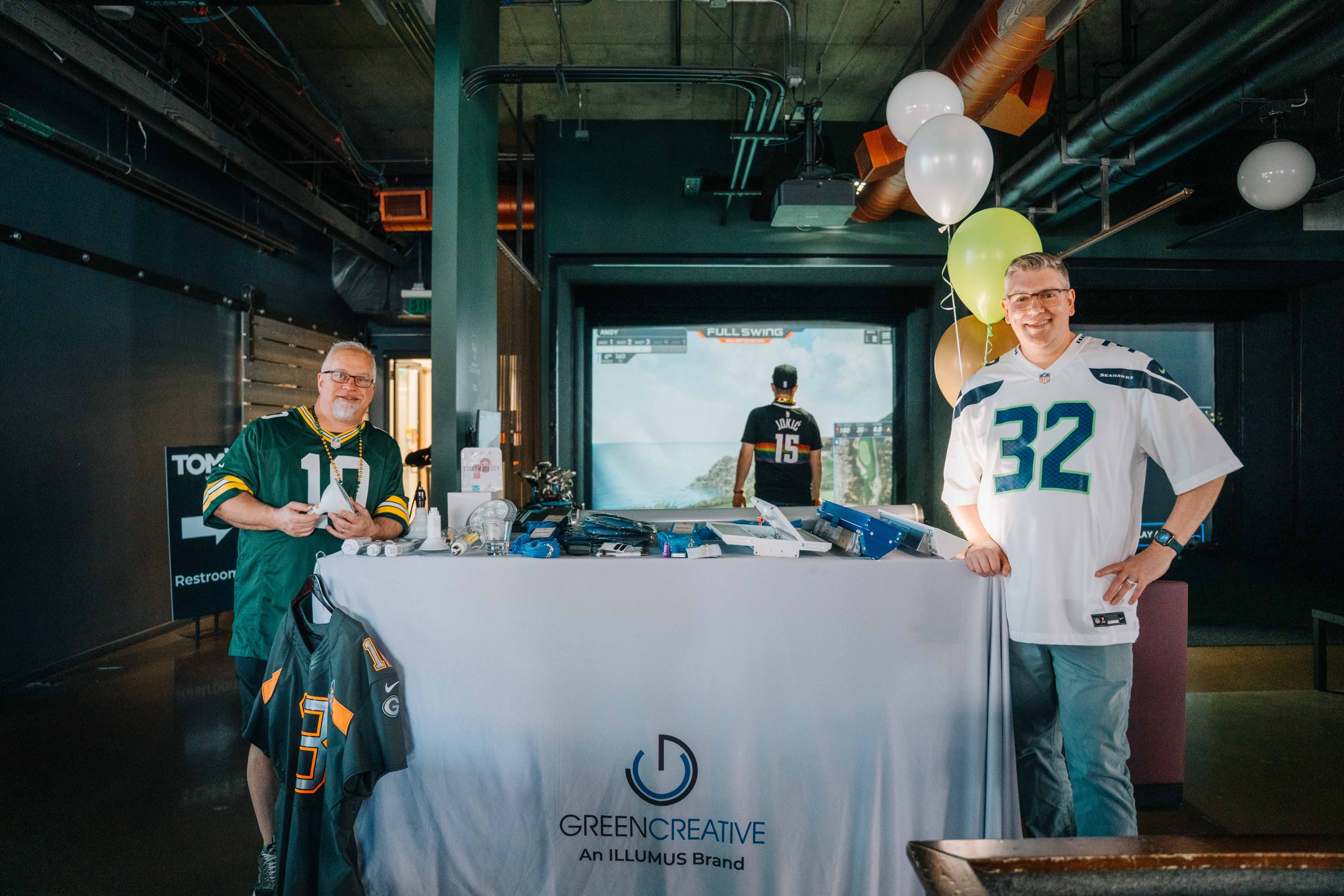 Two men stand behind a table with promotional materials and branded items at an indoor event, smiling for the photo. One wears a green sports jersey, and the other wears a white sports jersey with the number 32. Balloons are attached to a nearby pole