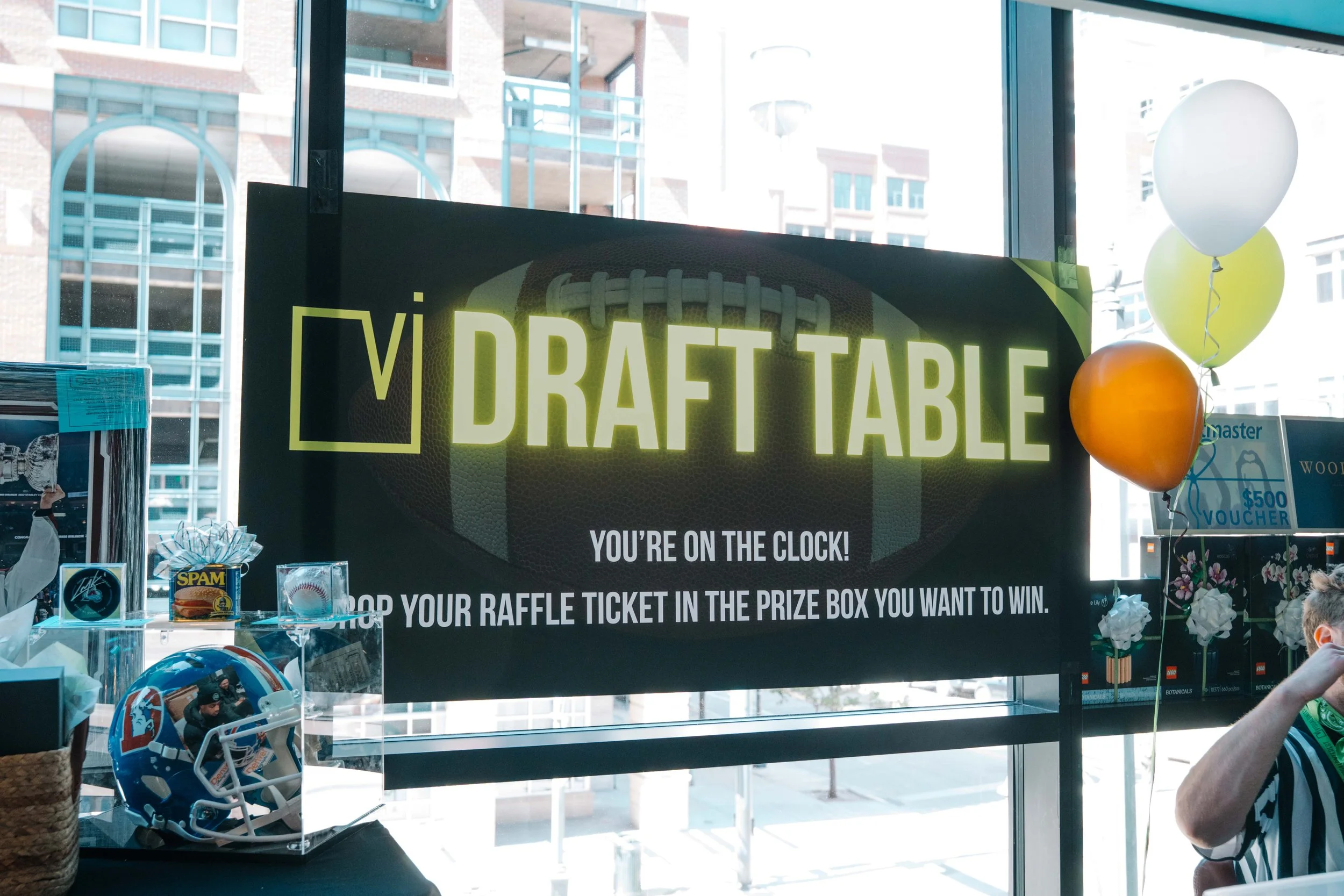 Sign reading 'VI Draft Table' with instructions to drop raffle ticket in prize box. Balloons tied to a counter, football helmet, and several prizes on display are visible in the background.