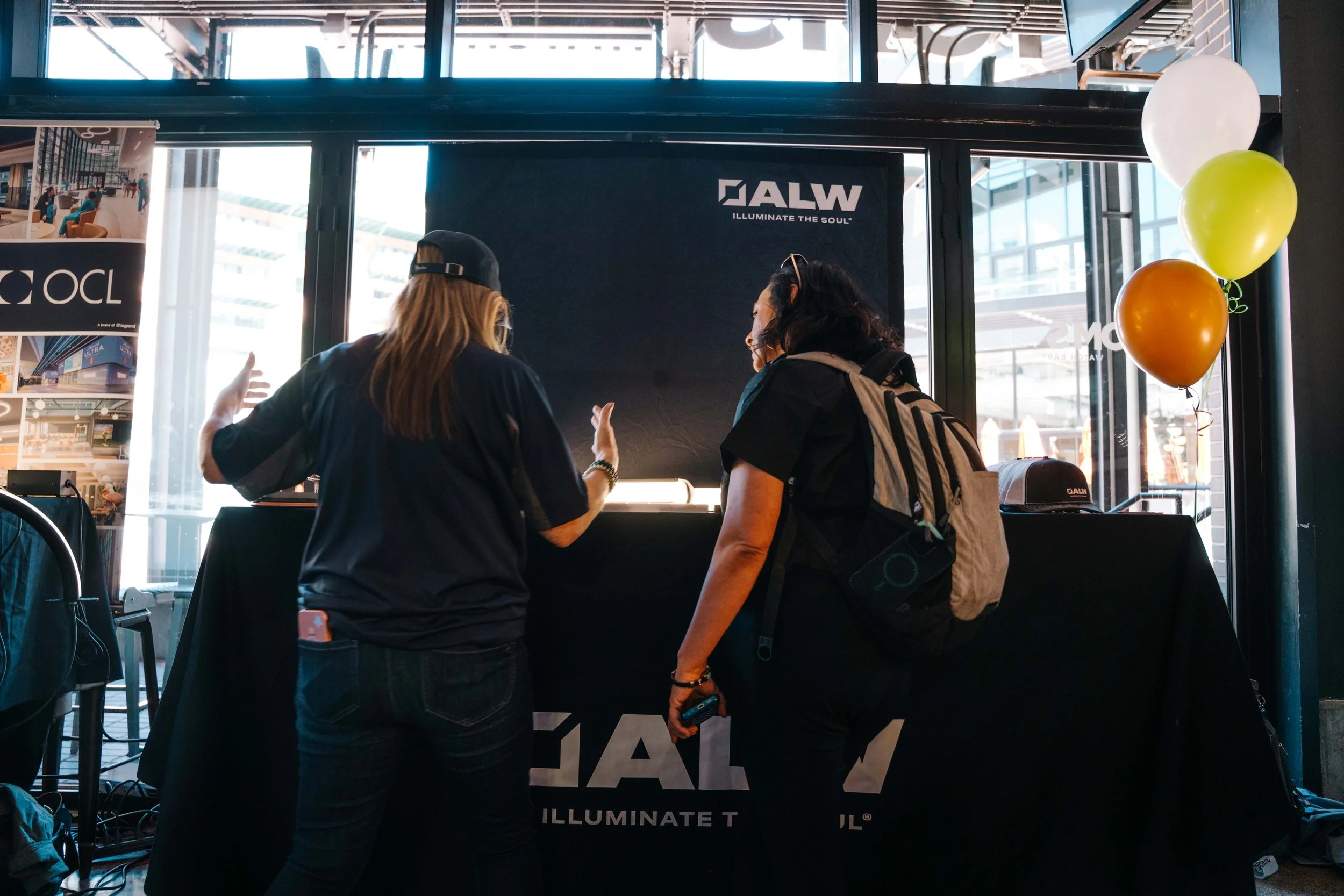 Two women are talking at an event booth with black banners displaying a logo and the slogan "Illuminate the Soul." One woman has blonde hair, a black cap backwards, and a black shirt, while the other has dark hair, glasses on top of her head, and a b