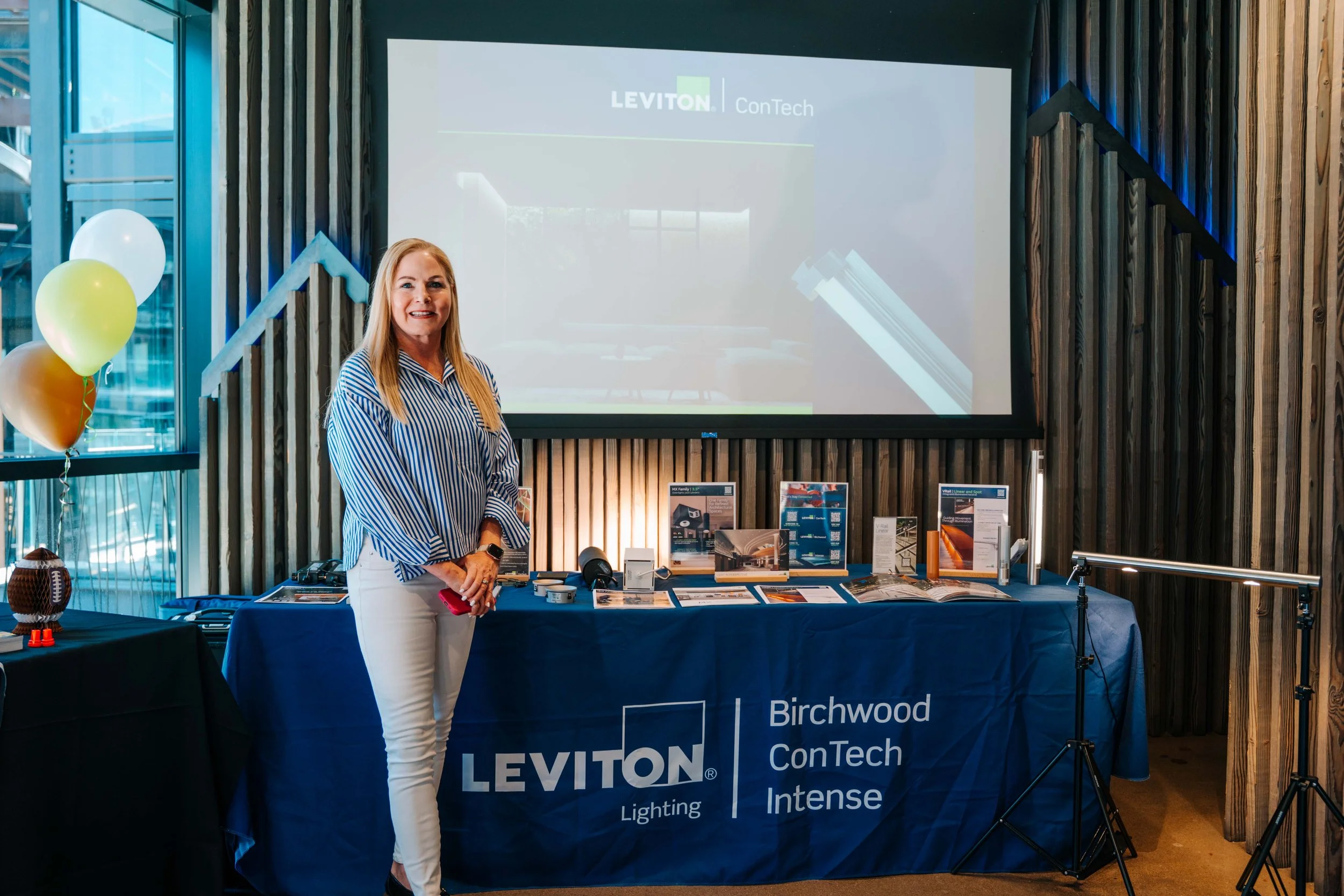 A woman with blonde hair, wearing a striped shirt and white pants, stands in front of a table at a corporate event. The table displays brochures and has a banner with the text 'Leviton' and other related information. Behind her, a large screen shows 