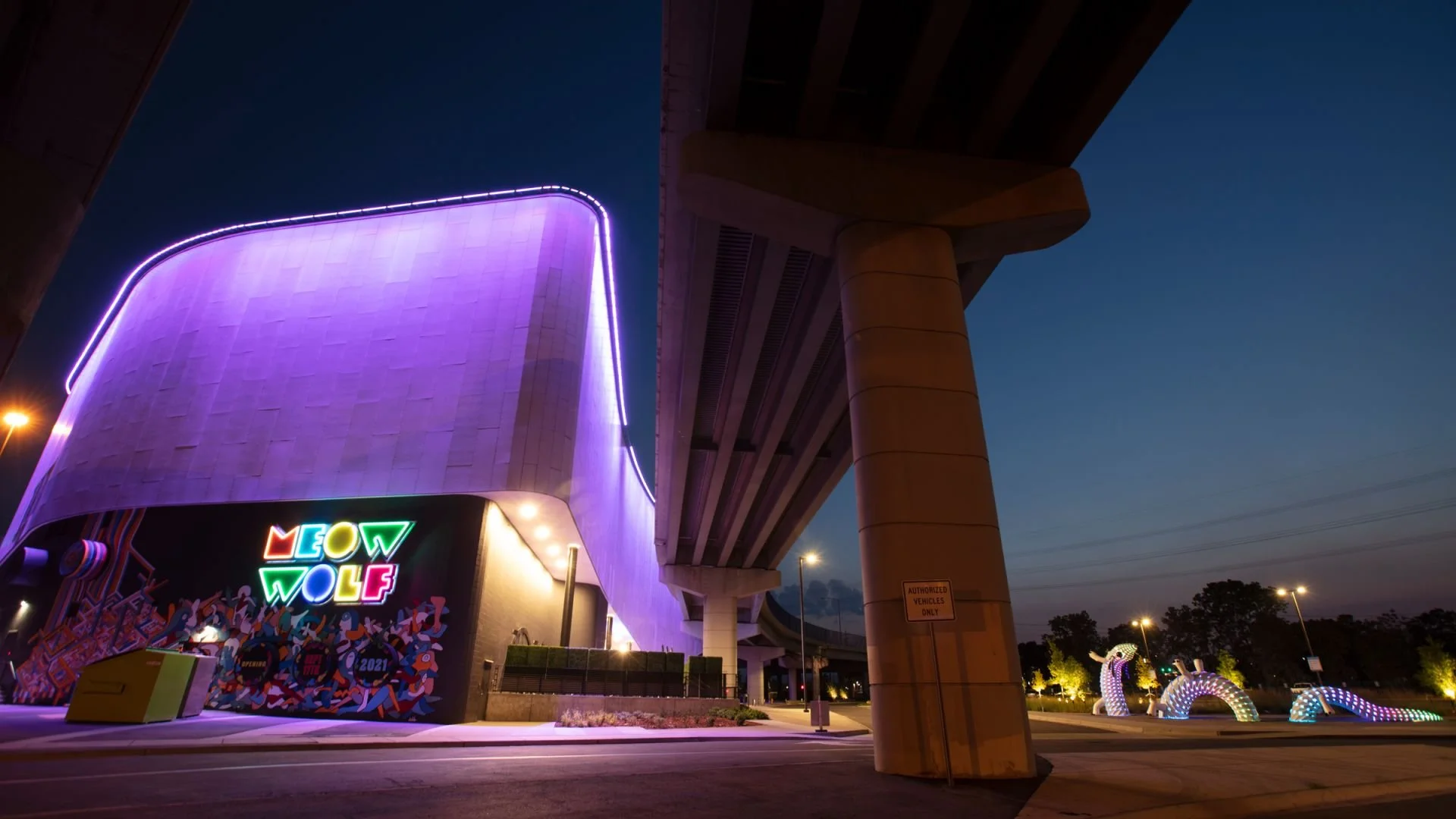 Modern building with vibrant colorful neon lights that spell 'MEOW WOLF,' under an overpass at dusk, with lit-up rainbow and unicorn sculptures in the background.