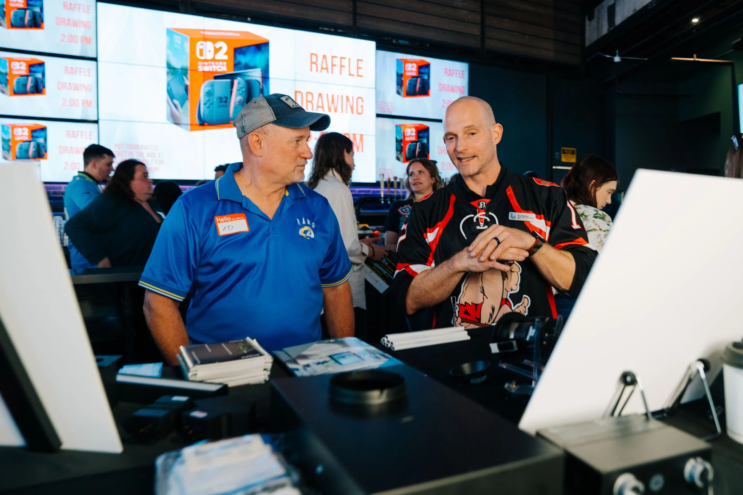 At a raffle drawing event, two men appear to be in conversation. The man on the left wears a blue shirt with a name tag, and the man on the right wears a black hockey jersey. They are standing behind a table with pamphlets and other items. In the bac