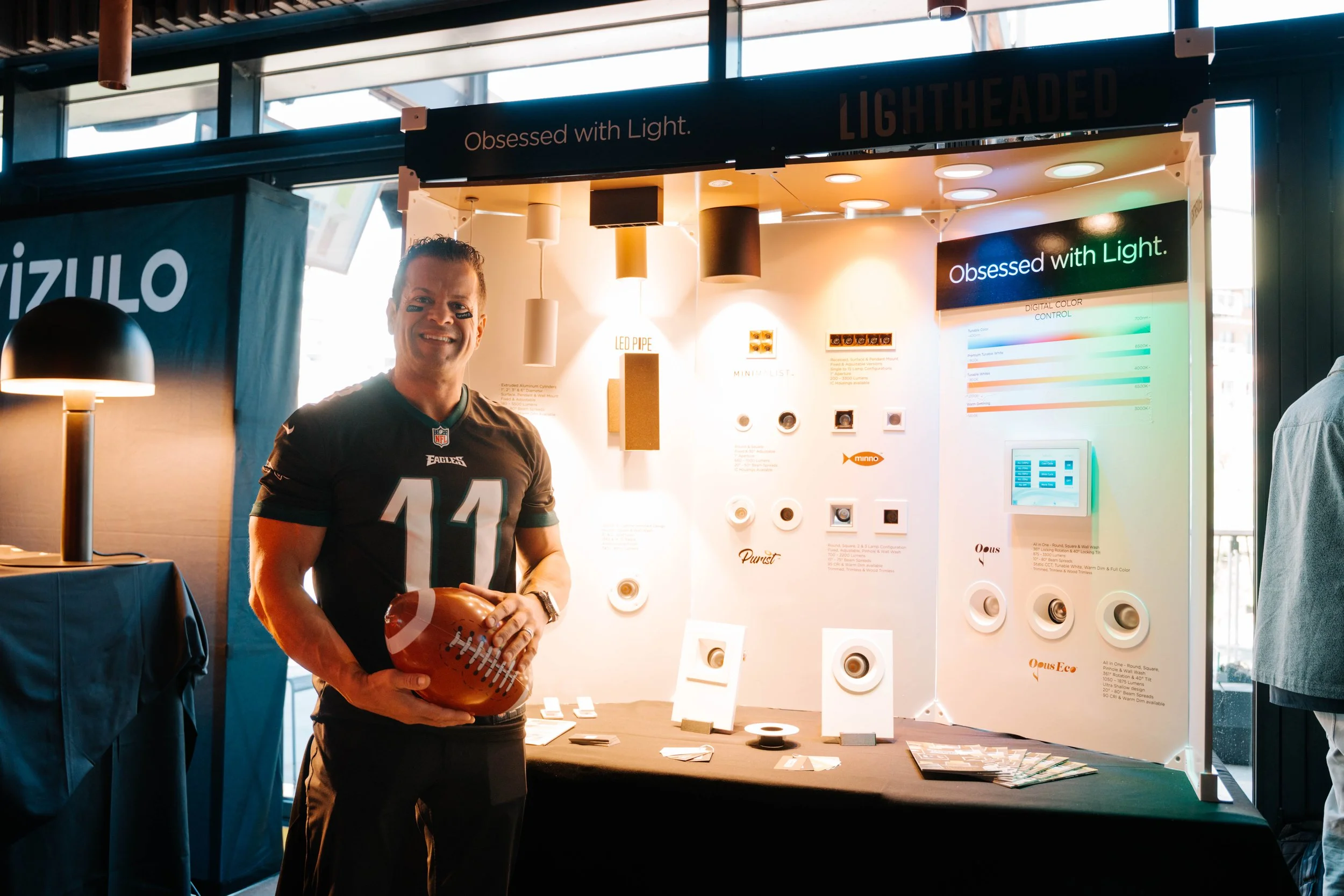 A man wearing a football jersey holding an orange football stands in front of a display booth labeled 'Lightheaded.' The booth showcases various lighting fixtures and control systems, illuminated with warm lighting.