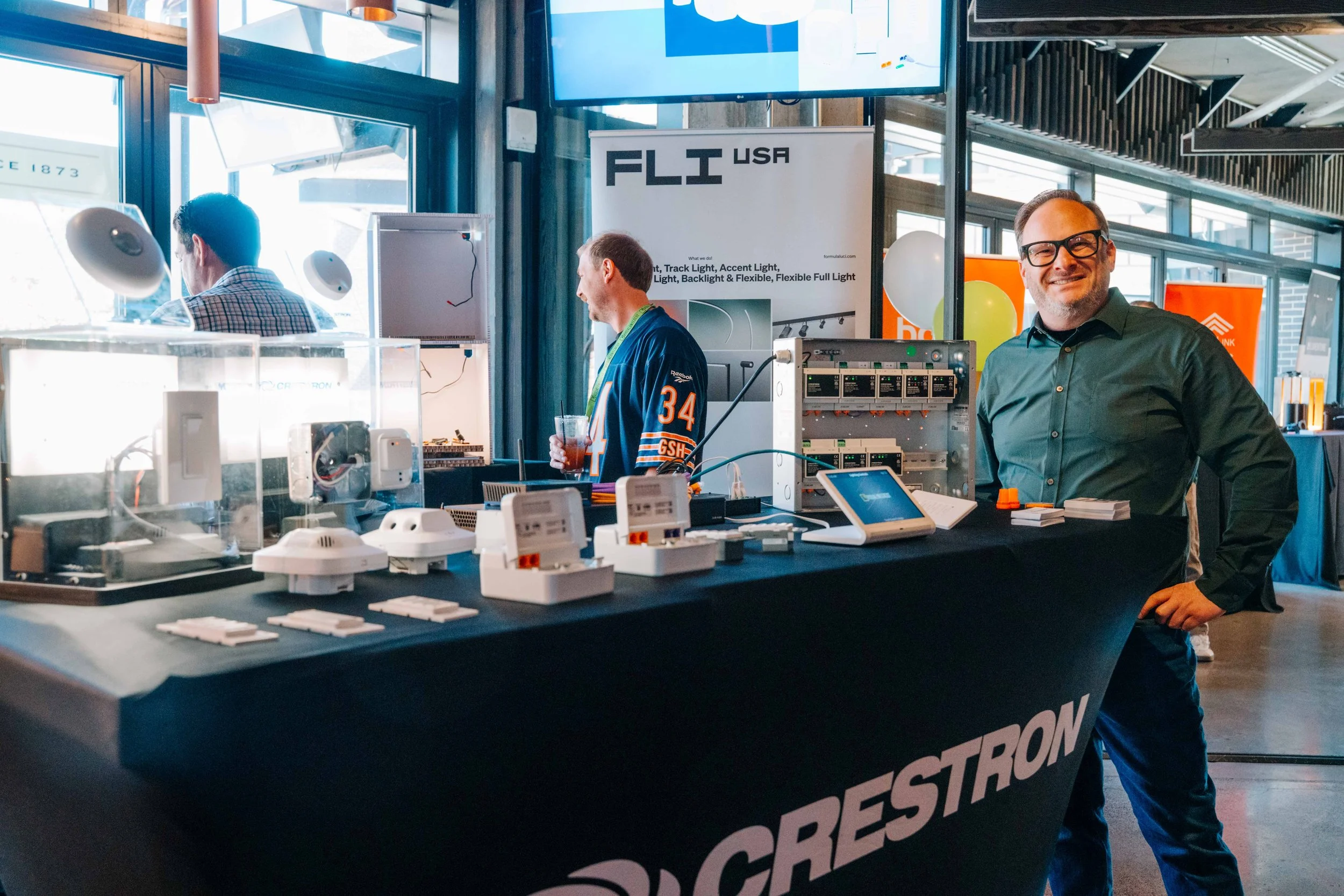 A man with glasses and a green shirt smiling at a tech exhibition booth with electronic components on display and a sign reading 'FLI USA'.