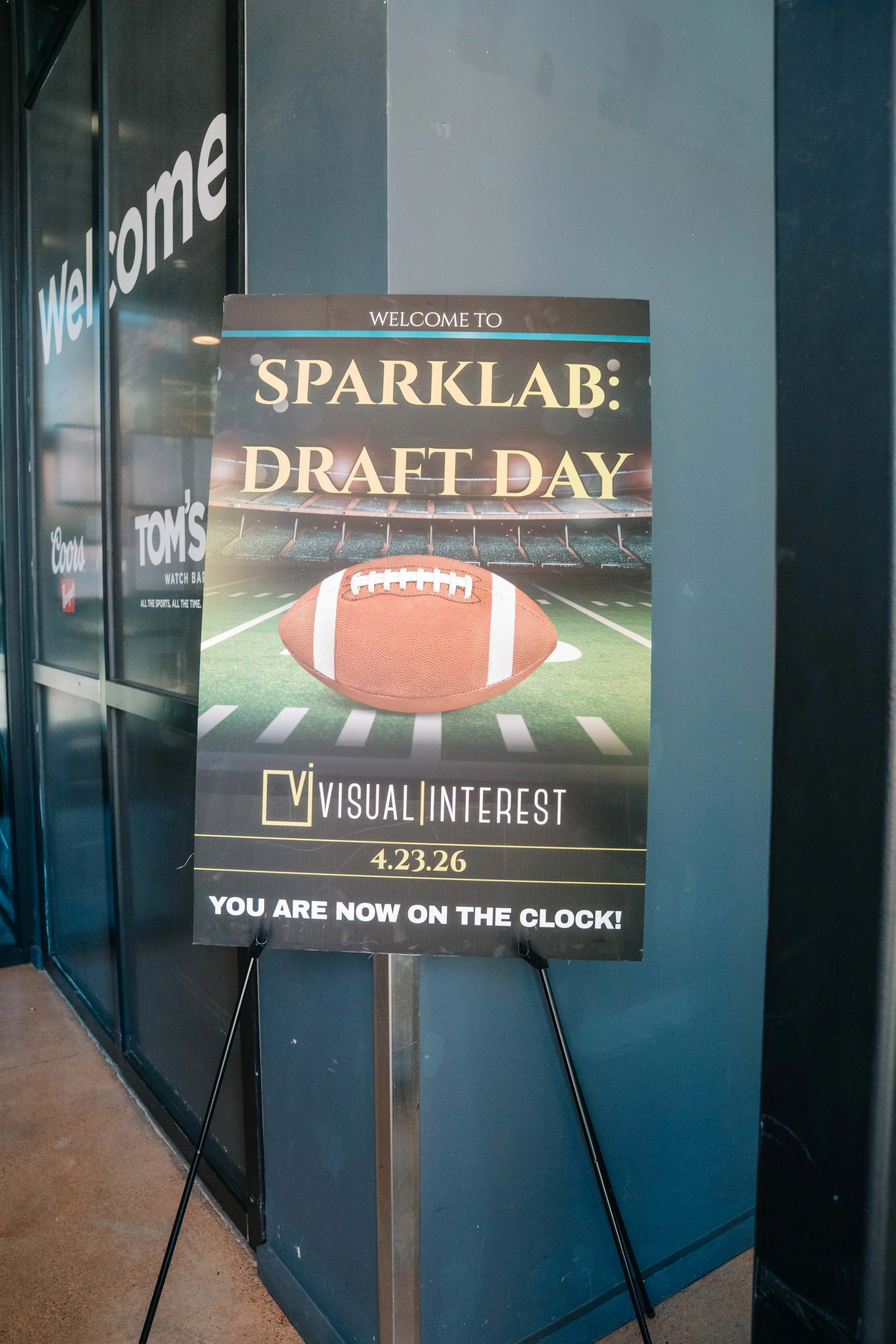 A sign on an easel near a stadium features football imagery and announces SparkLab Draft Day event on April 23, 2026, at 2:00 PM, indicating the viewer is now on the clock for the event.