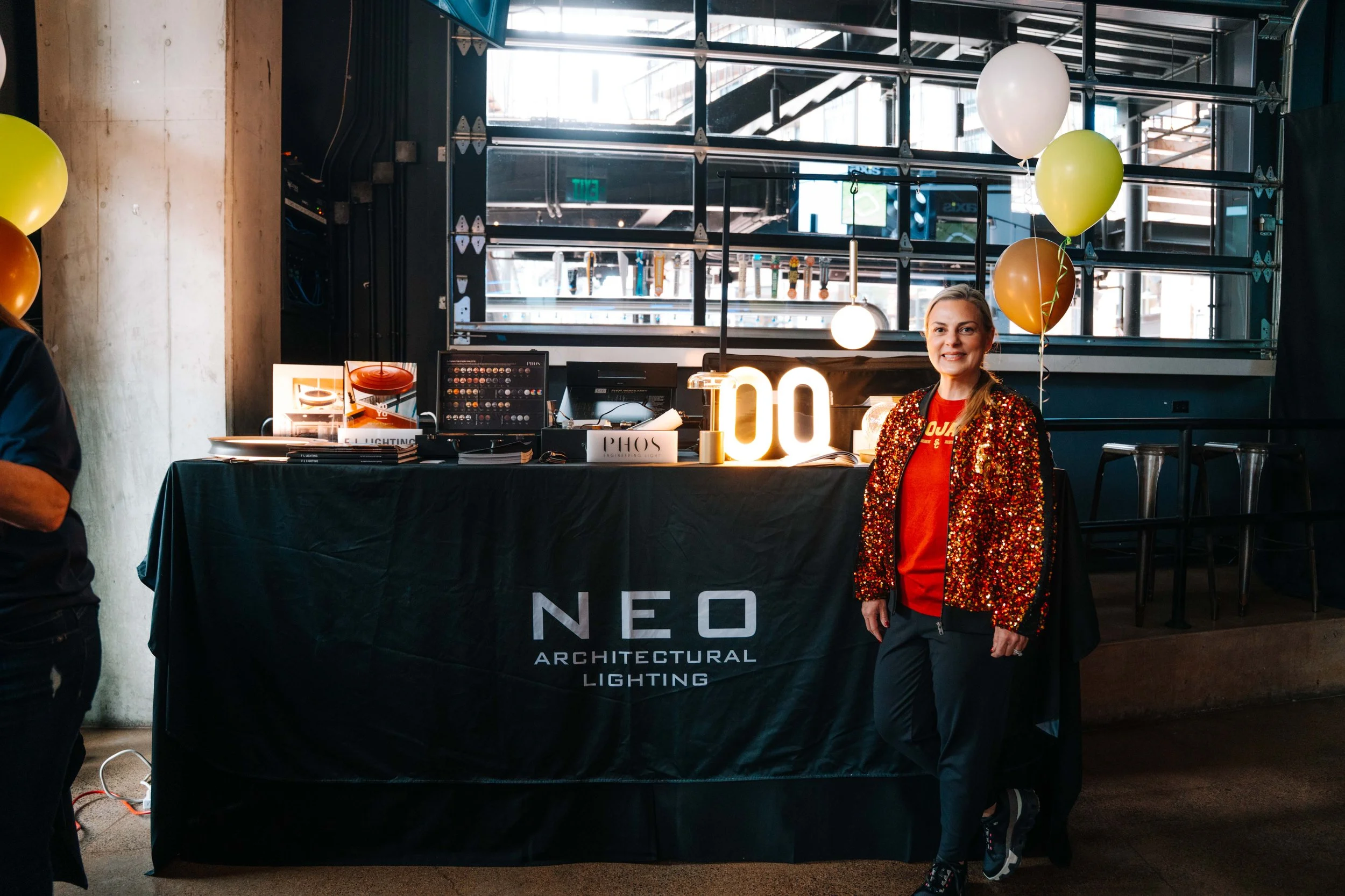 A woman in a red shirt and a red sequin jacket standing next to a table with a sign that reads "NEO ARCHITECTURAL LIGHTING", balloons, and a lamp. The background shows large windows and industrial-style interior with stairs and structures.