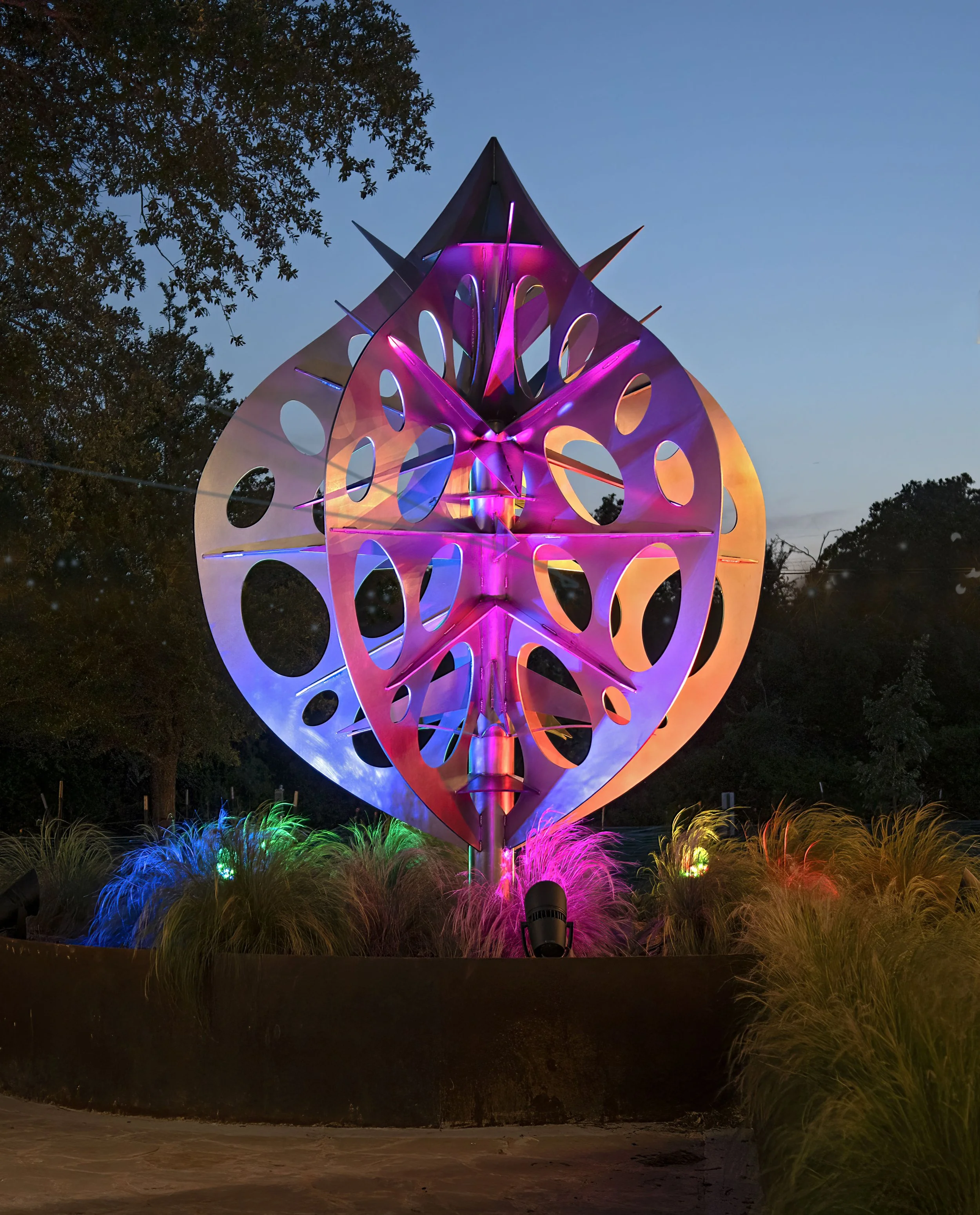 Colorful abstract outdoor sculpture illuminated with pink, purple, and blue lights at dusk, surrounded by ornamental grasses and trees.