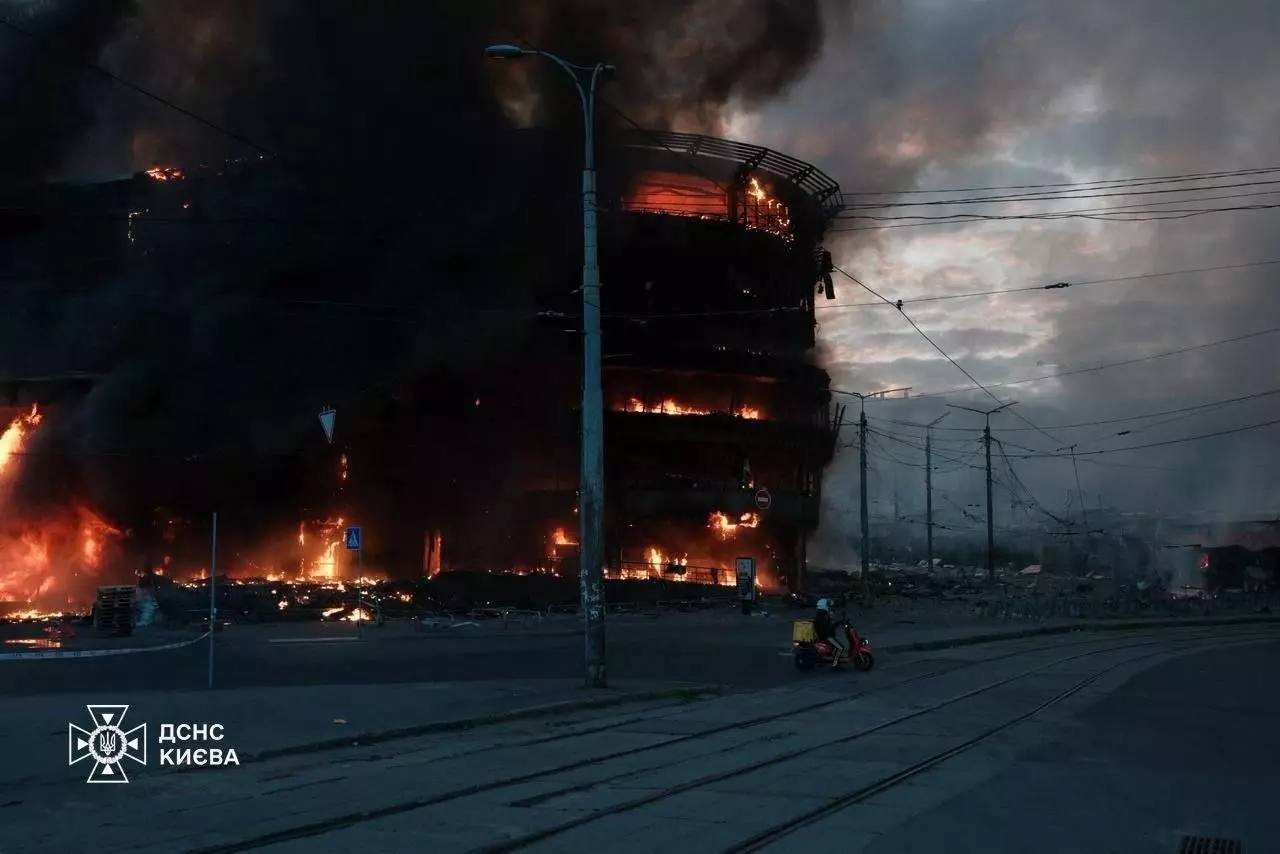 A multi-story building is on fire with flames and thick black smoke billowing out. The fire has caused significant damage, with visible destruction on the ground level. A motorcyclist rides past the scene on a nearby street, and utility poles with power lines are present. The image appears to be taken during dusk or dawn. There is Ukrainian text and logo of the State Emergency Service of Kyiv in the bottom left corner.