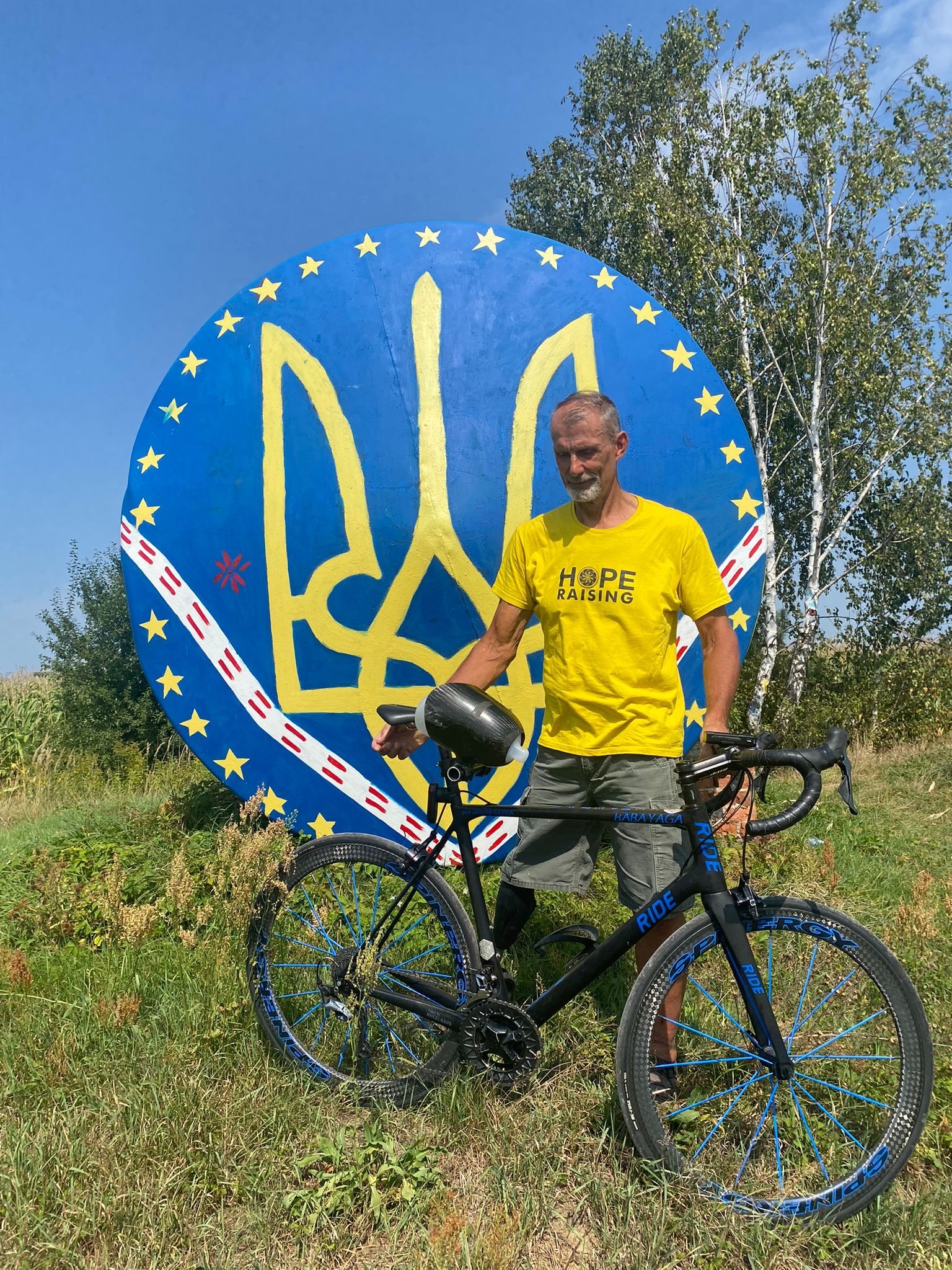 A man wearing a yellow T-shirt with the words 'HOPE RAISING' and a peace symbol, standing outdoors with a black bicycle that has blue accents. Behind him is a large circular sign with a yellow trident symbol surrounded by yellow stars on a blue background, and a tree with green leaves and a bright blue sky.