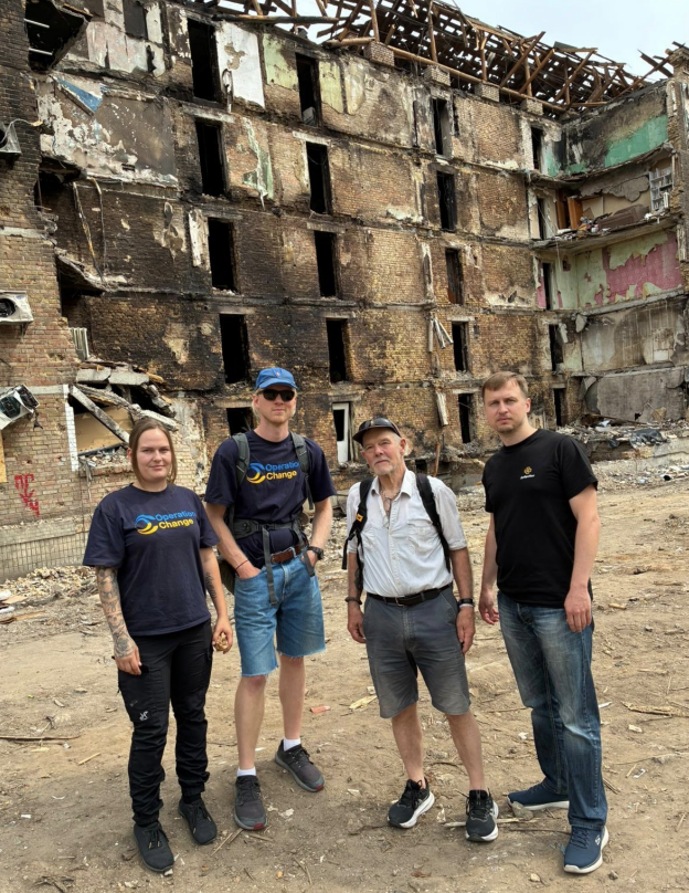 Four people standing in front of a destroyed building, with exposed brick walls and collapsed roof.