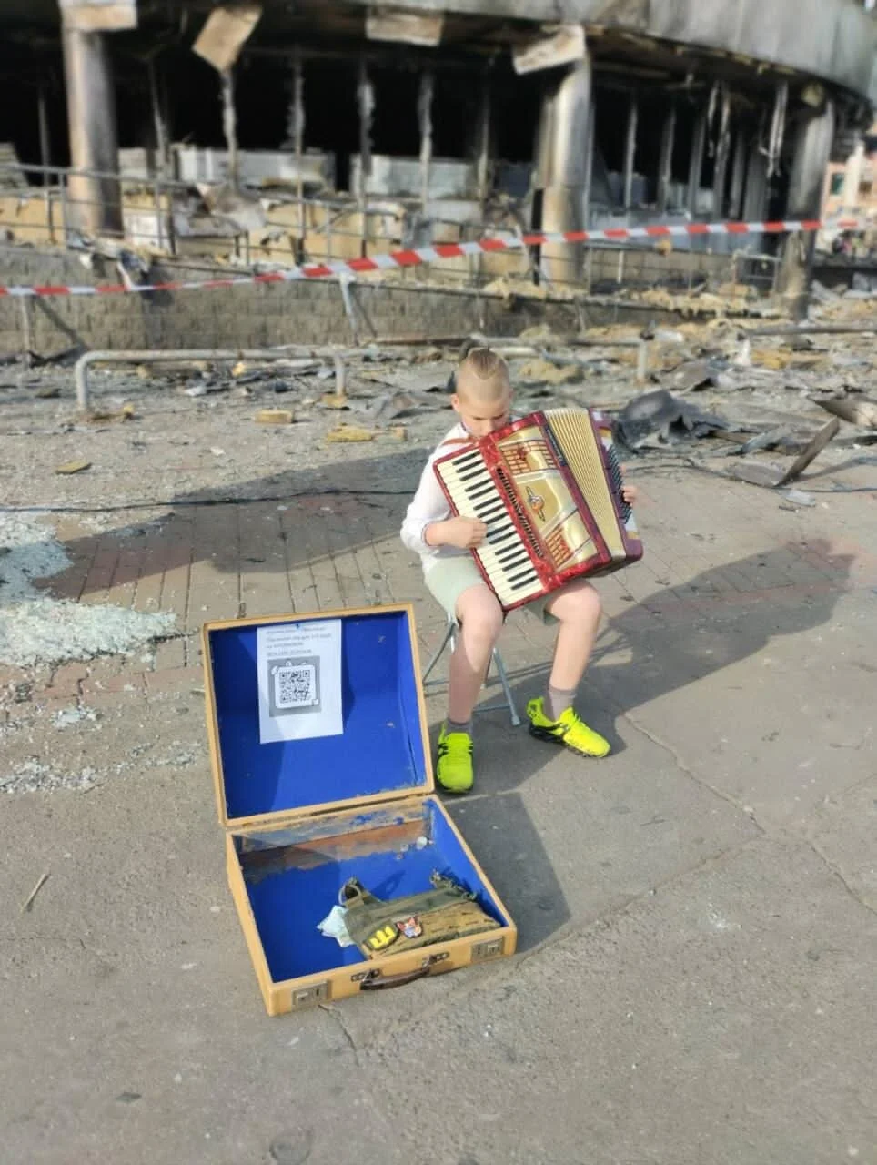 A young boy sitting on a small chair, playing an accordion, amid the ruins of a burned building. An open suitcase with a toy gun and other items is on the ground nearby, in front of him.
