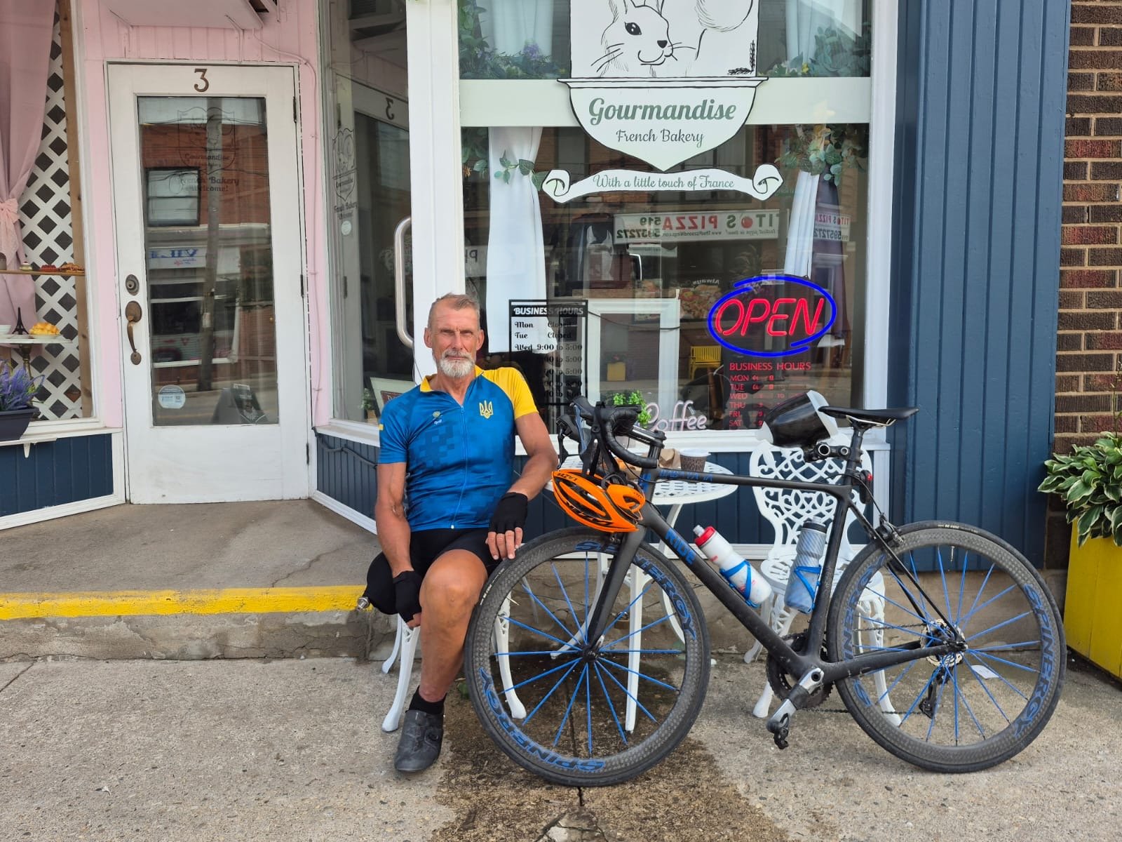 A man in cycling gear seated outside a bakery named Gourmandise French Bakery, with a black mountain bike in front of him. The bakery has a large window with a neon 'OPEN' sign and a sign for business hours.