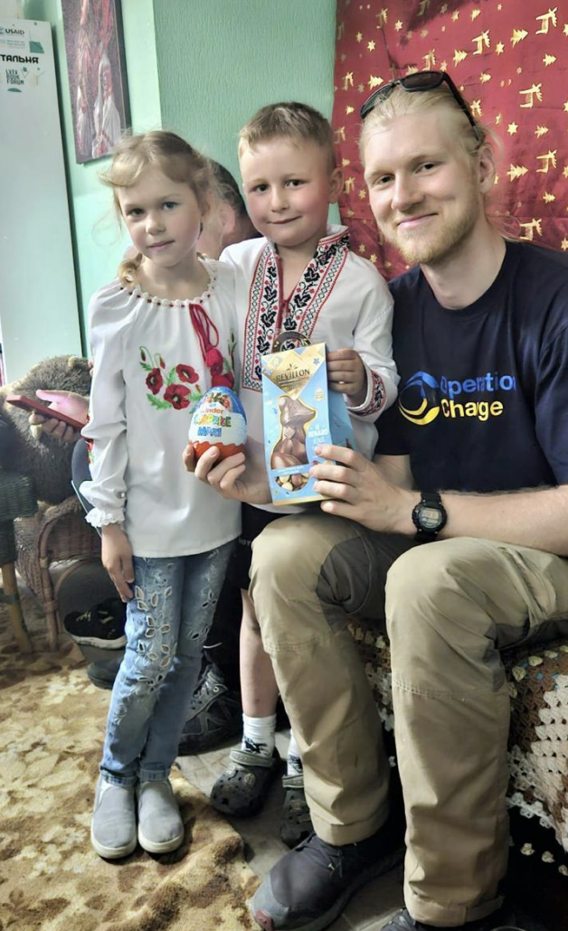 Three children, two girls and a boy, and a young man sitting on a couch in a room decorated with green walls, a red curtain with gold stars, and a patterned carpet. The children are holding Easter chocolates and are dressed casually, with the girl wearing a white embroidered blouse and the boy wearing a traditional embroidered shirt. The young man is wearing a black T-shirt with yellow and blue text, khaki pants, and sunglasses on his head.