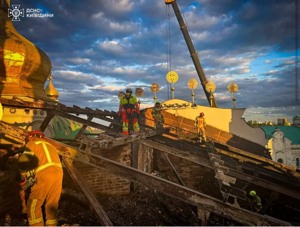 Firefighters working on the ruins of a collapsed or damaged structure with debris, construction equipment, and a partly damaged roof. The scene is at sunset or sunrise with a sky full of clouds.