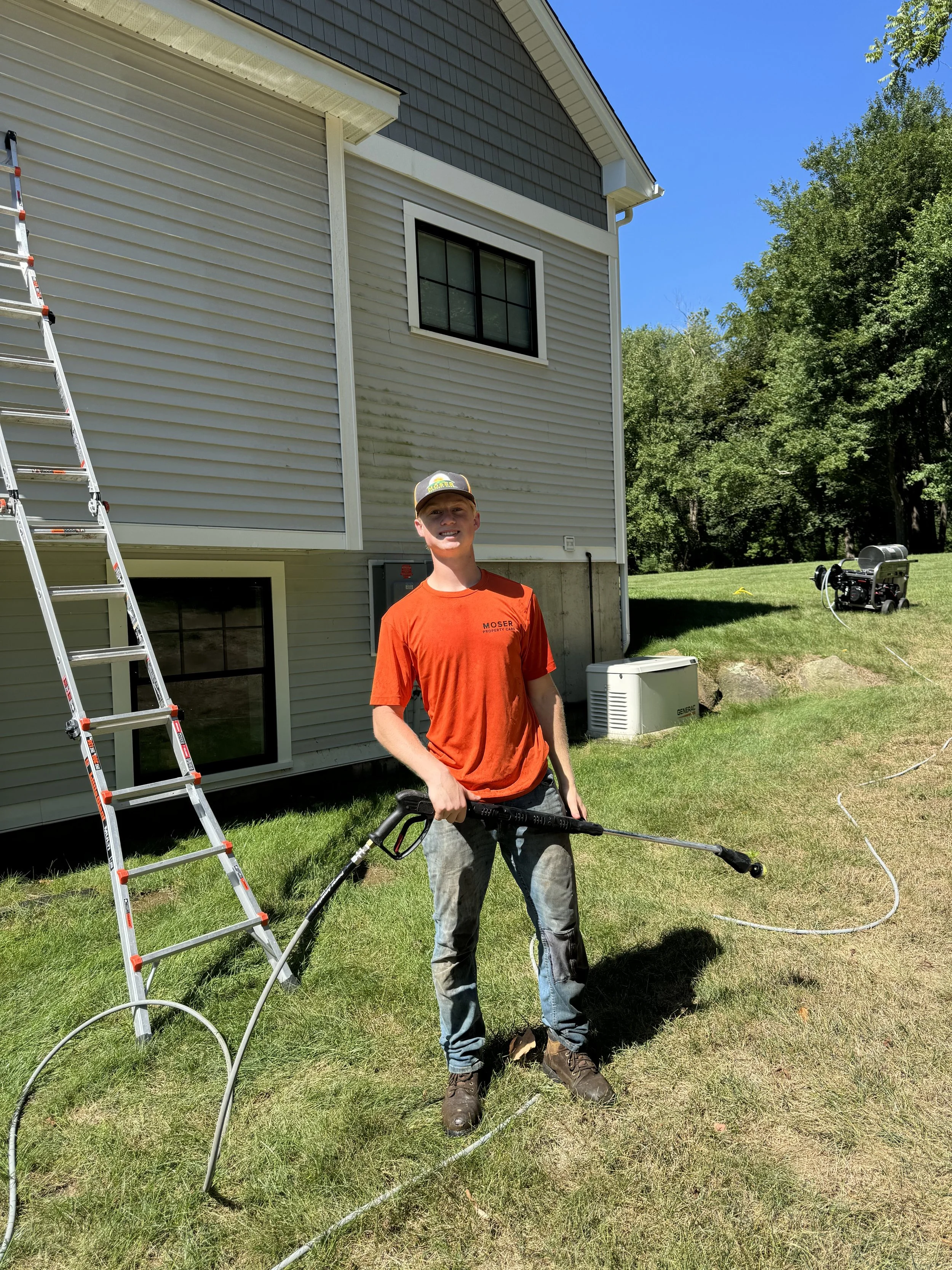 A young man wearing an orange t-shirt and jeans holding a pressure washer wand stands on a grassy yard next to a house. There is a ladder leaning against the house, and some equipment can be seen on the lawn, likely for cleaning or maintenance.