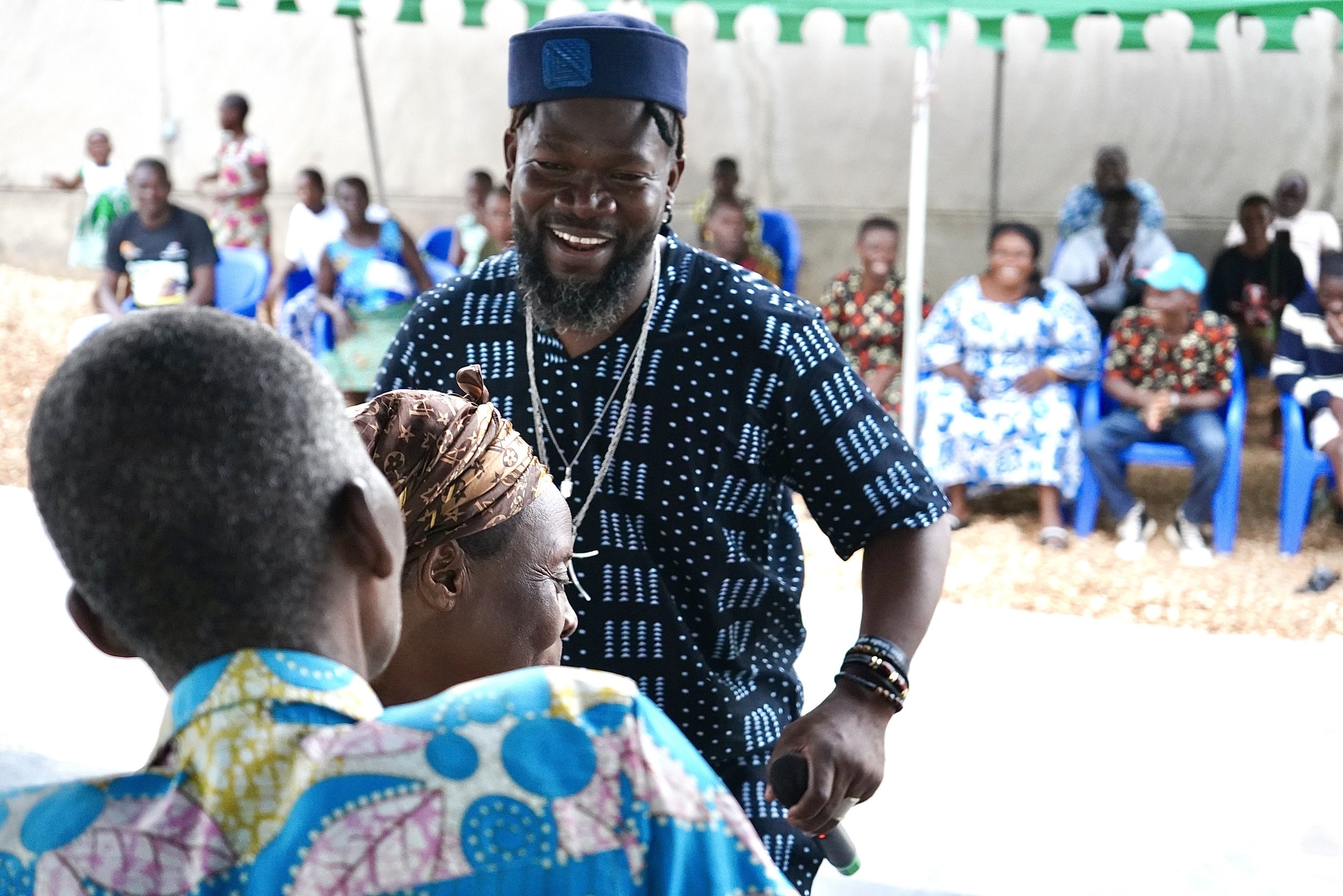 Un homme souriant avec une barbe et un chapeau bleu remet un prix ou contribue à une cérémonie, entouré de plusieurs personnes assises en arrière-plan lors d'un événement en plein air.
