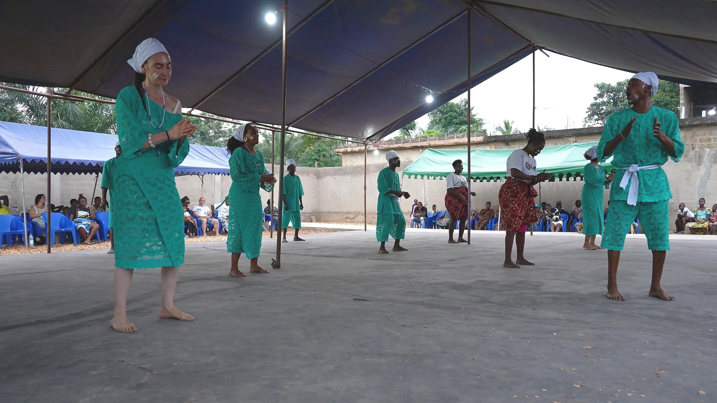 Performance de théâtre ou danse traditionnelle sous une tente, avec des personnes portant des vêtements en tissu turquoise et d'autres en vêtements rouges et blancs, avec un public assis en arrière-plan.