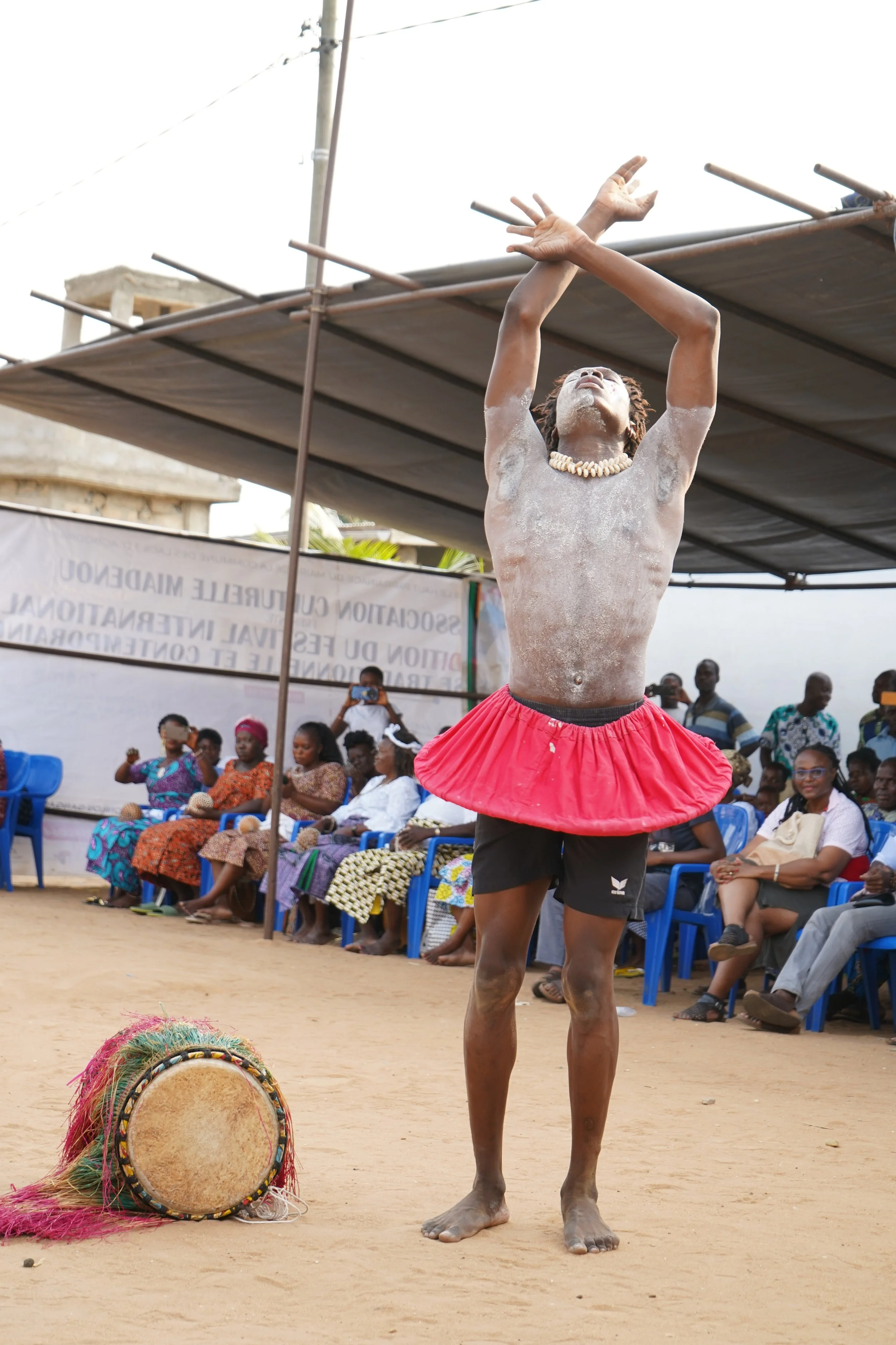Un homme danseur traditionnel en costume, portant un collier de coquillages, un pagne rouge et un short noir, lors d'une performance dans un espace en plein air avec un public assis.