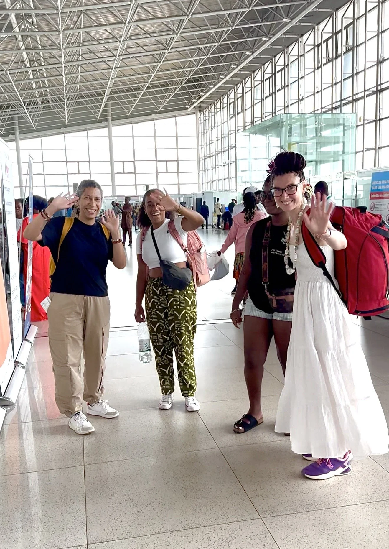 quatre femmes souriantes, portant des sacs à dos, saluant à l'aéroport, à l'intérieur, avec une architecture en verre et acier en arrière-plan.
