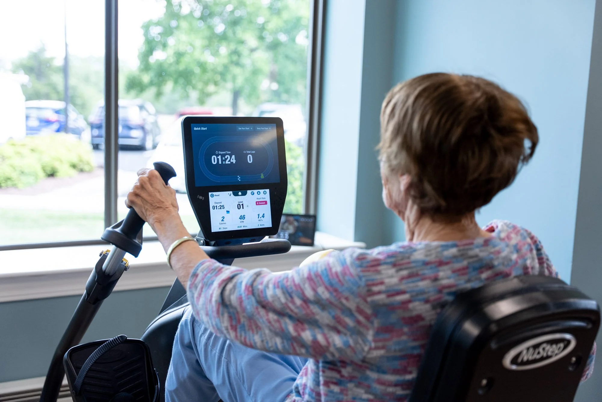 An older woman exercises on a seated rehabilitation bike while viewing her activity results on its digital screen.