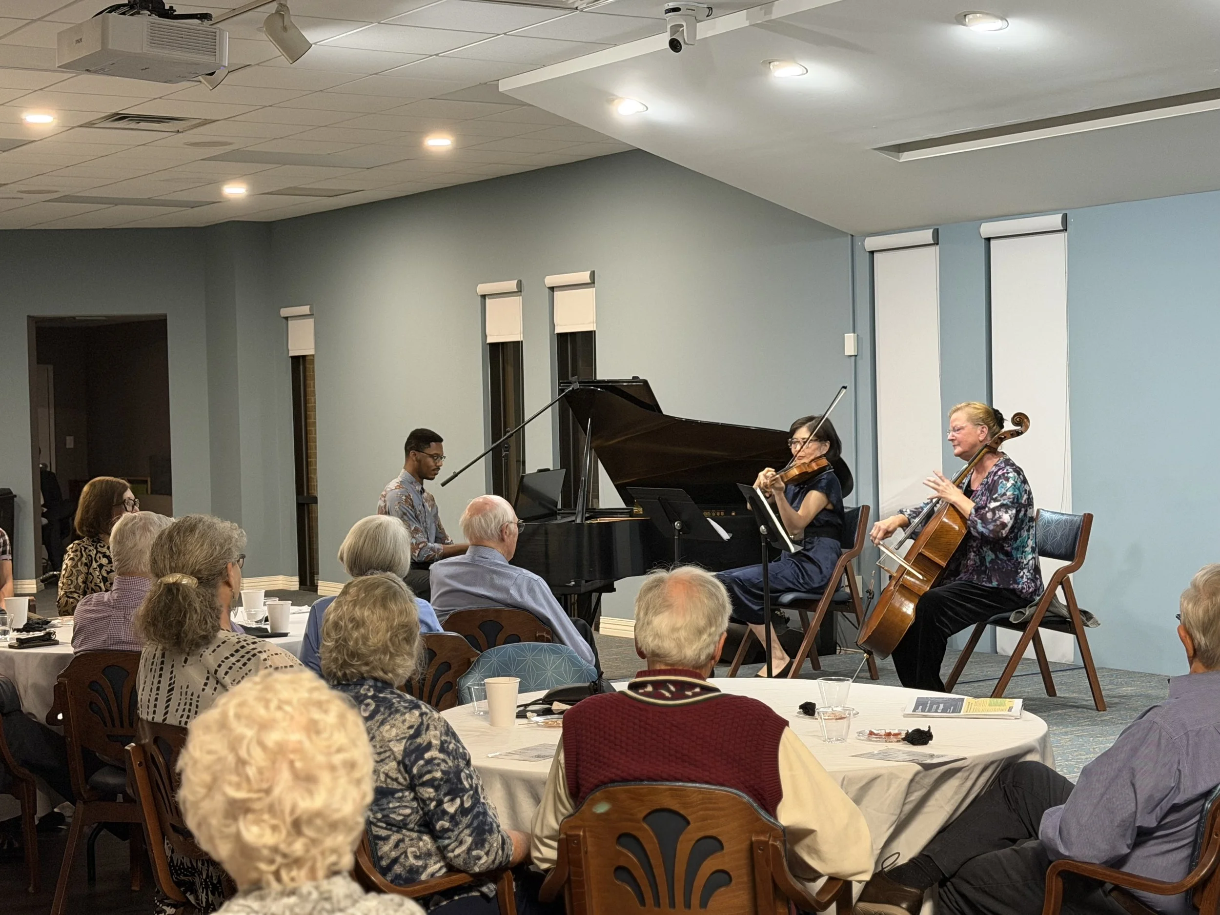 a group of people watching three individuals play cello, violin, and piano