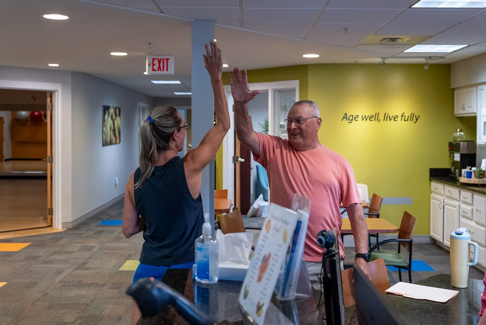 VMRC Wellness Center staff and member high-fiving at the front desk beneath an "Age well, live fully" sign.