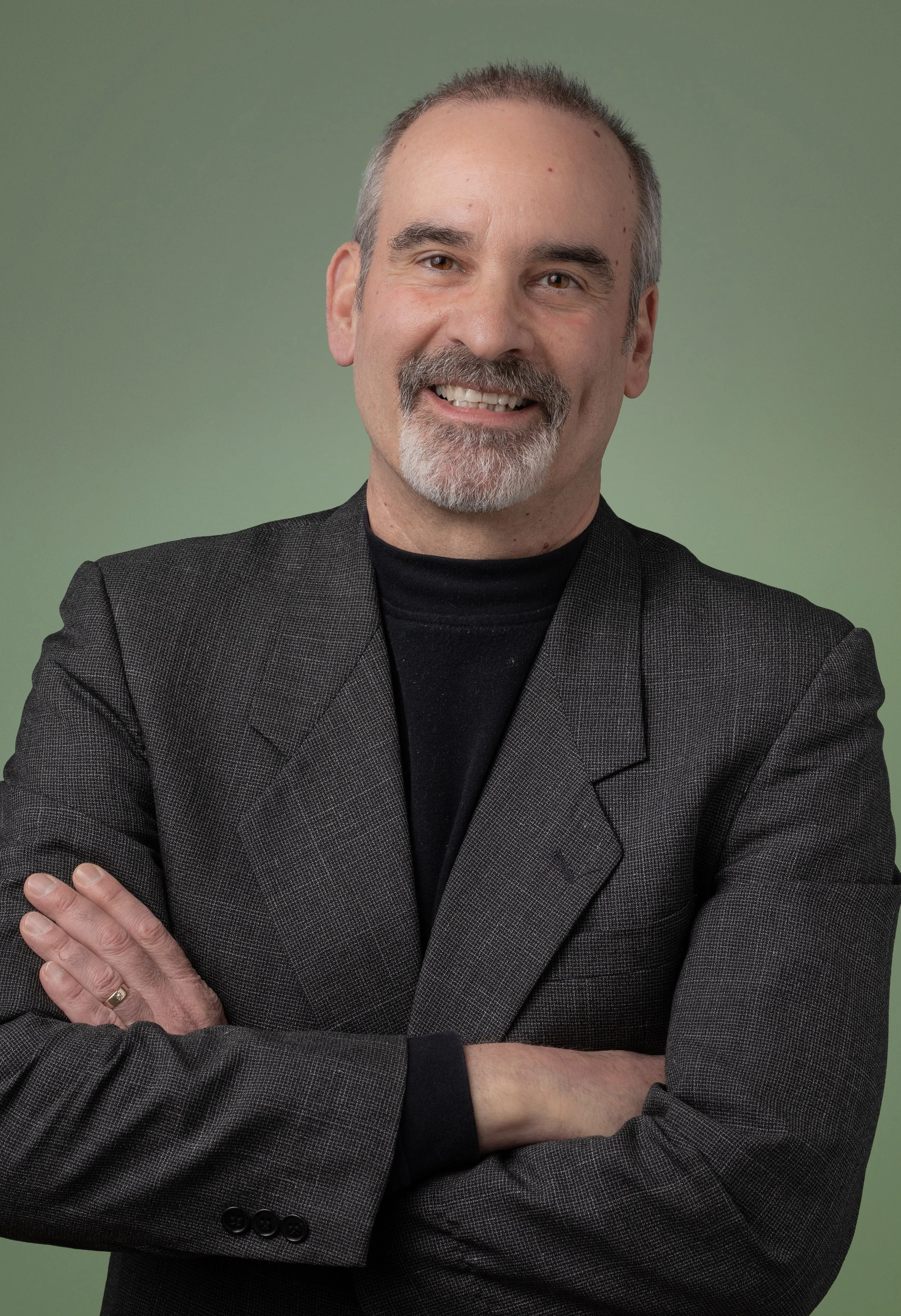 Headshot of a middle-aged man with gray hair, glasses, dressed in a black suit, white shirt, and patterned tie, smiling against a green background.