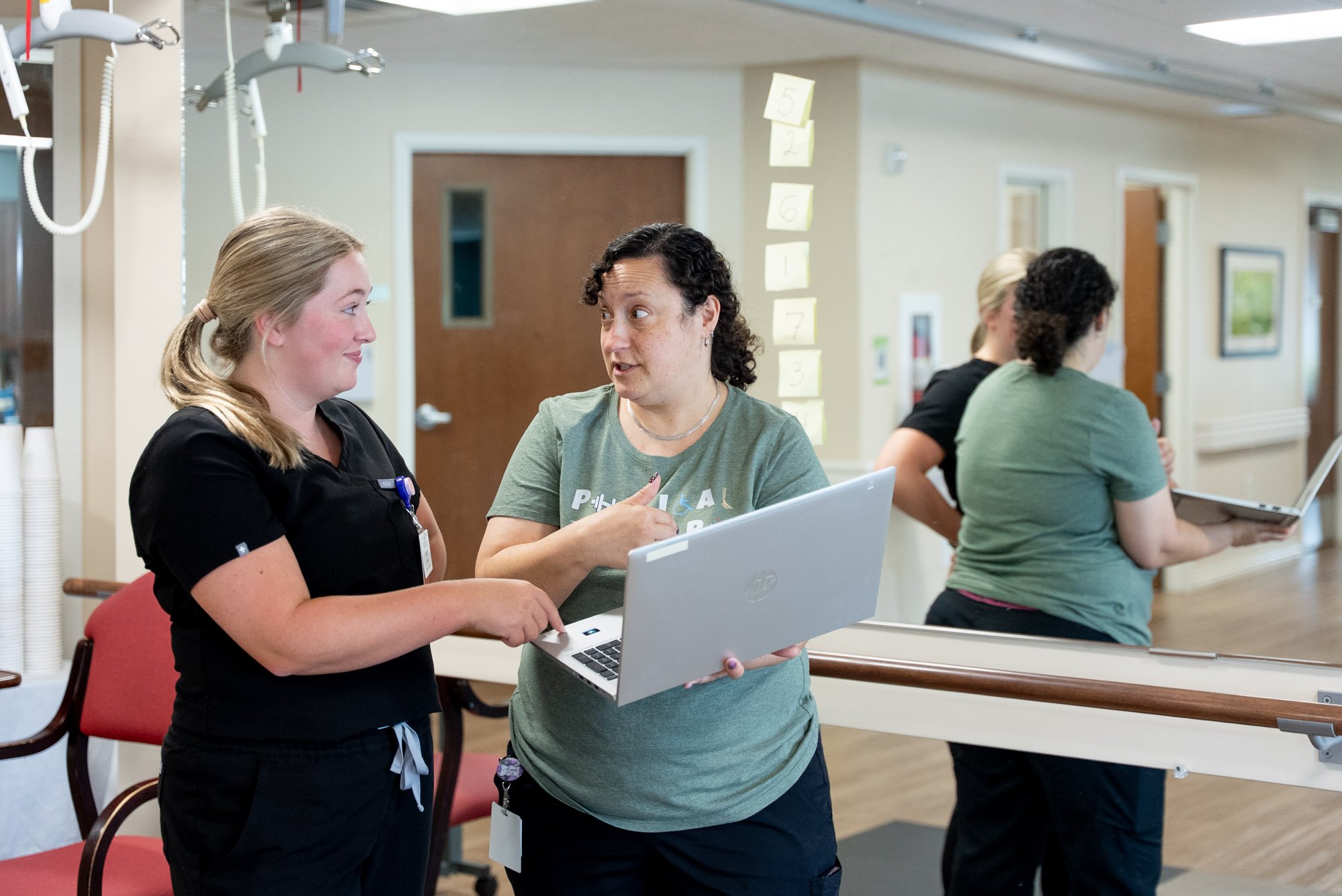 Two rehabilitation staff members review information on a laptop beside parallel bars in a therapy room.