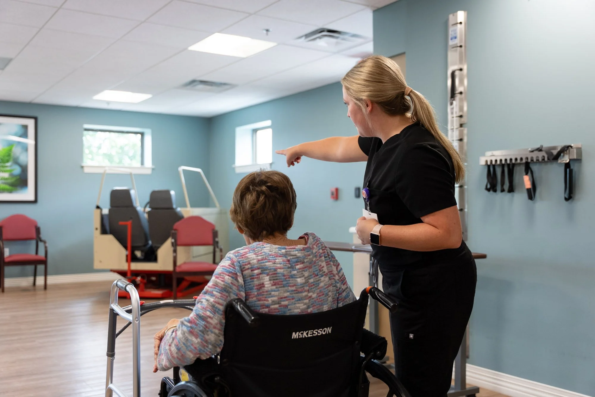 An occupational therapist points across a rehabilitation room while assisting an older woman seated in a wheelchair beside her walker.