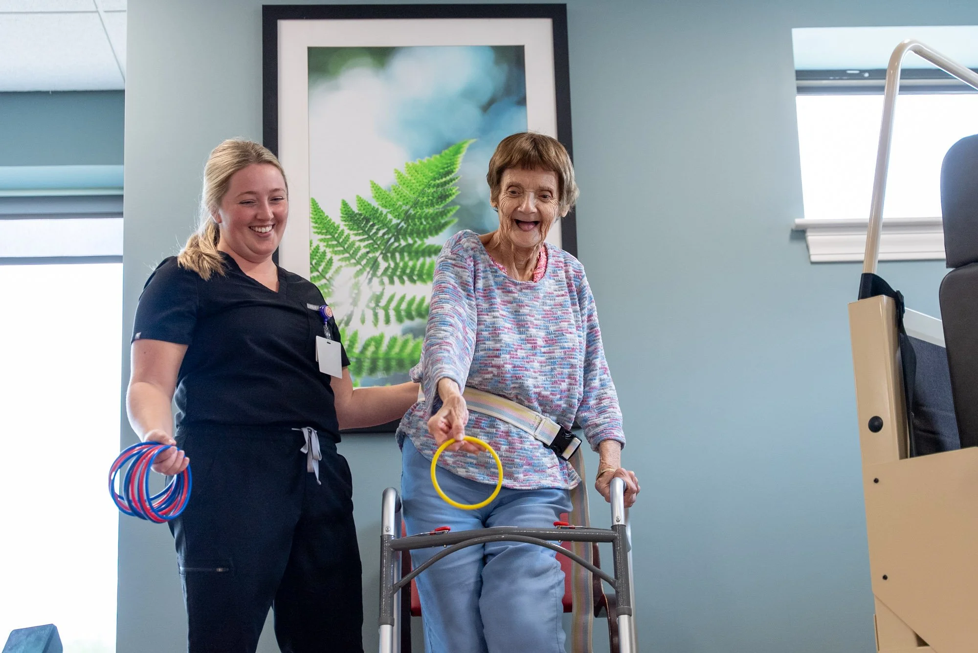 An occupational therapist smiles as an older woman uses a walker and tosses a yellow ring during a rehabilitation exercise.