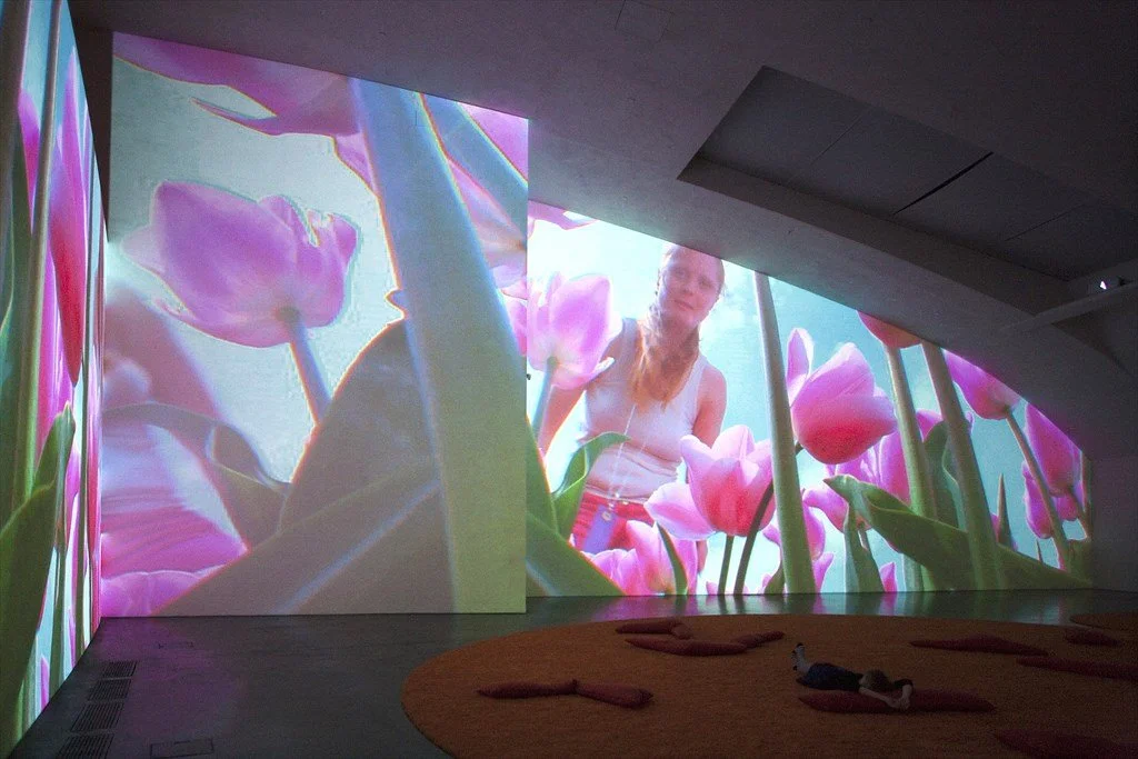 Indoor digital art installation projecting a large image of pink tulips and a woman with red hair in a white tank top among the flowers.